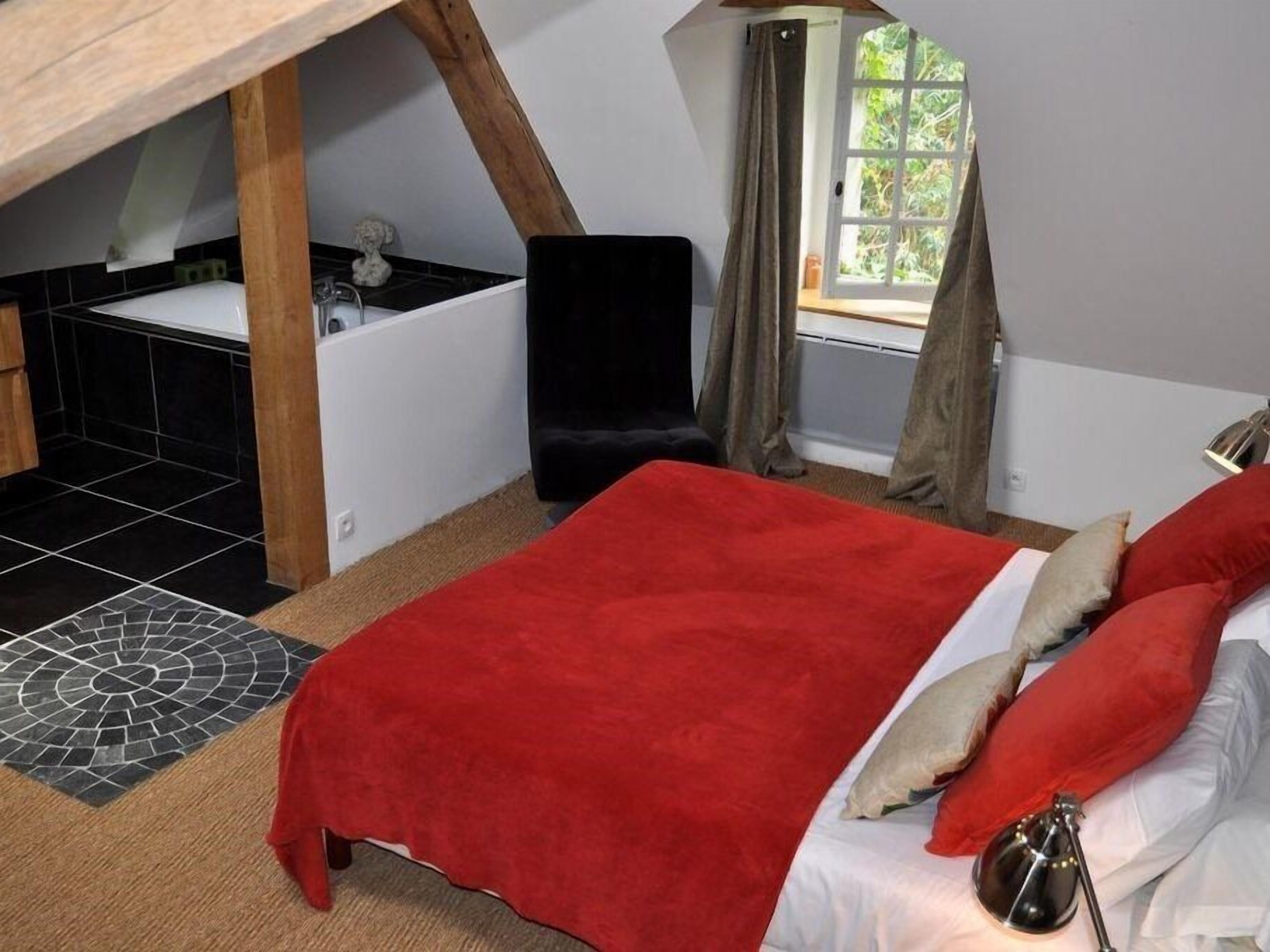 A bedroom with a sloped ceiling, a bed with red and white bedding, a black chair, a small sink, and a window with curtains.