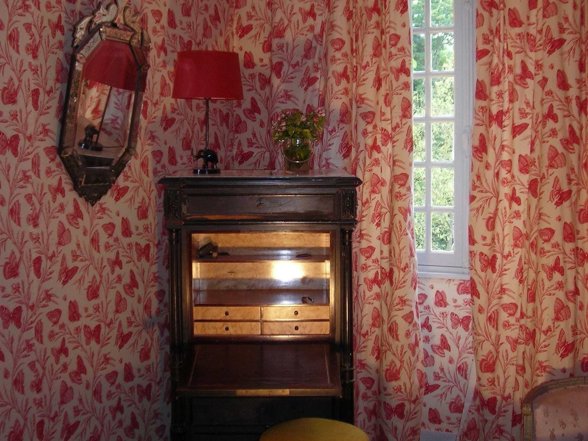 Quarto com papel de parede floral, um armário de madeira com gavetas, um abajur vermelho e uma janela com cortinas.