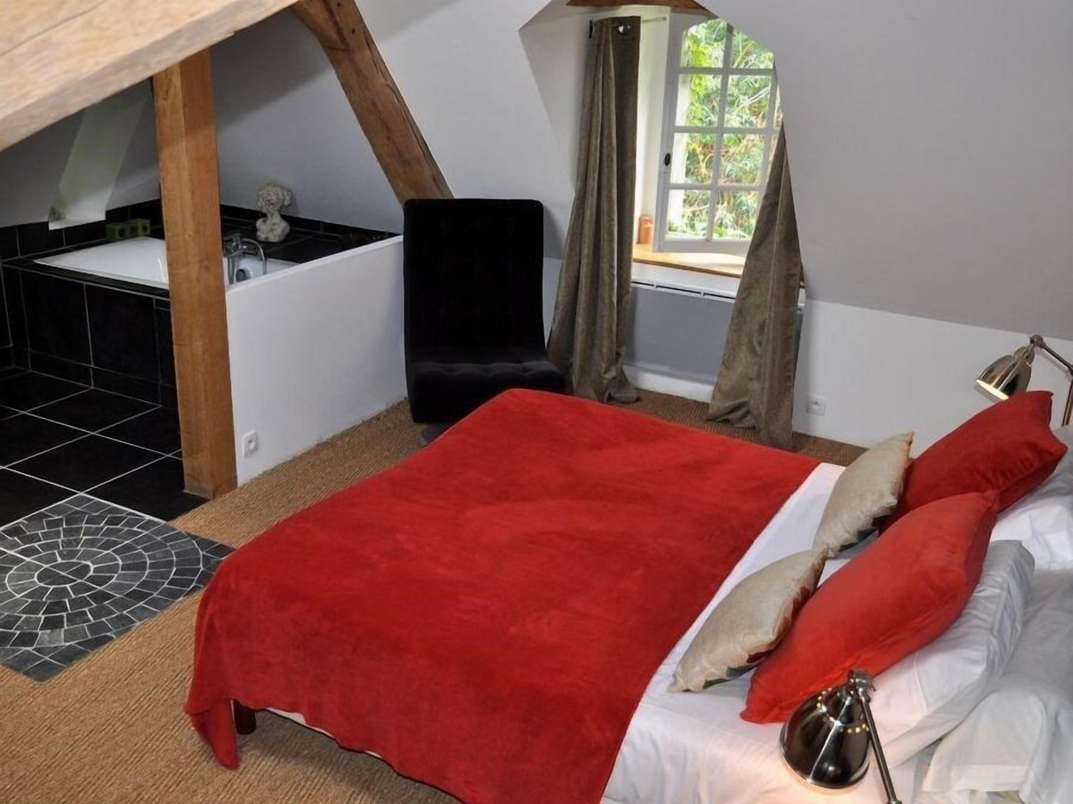 A bedroom with a red bedspread, white pillows, a black chair, and a window with curtains.