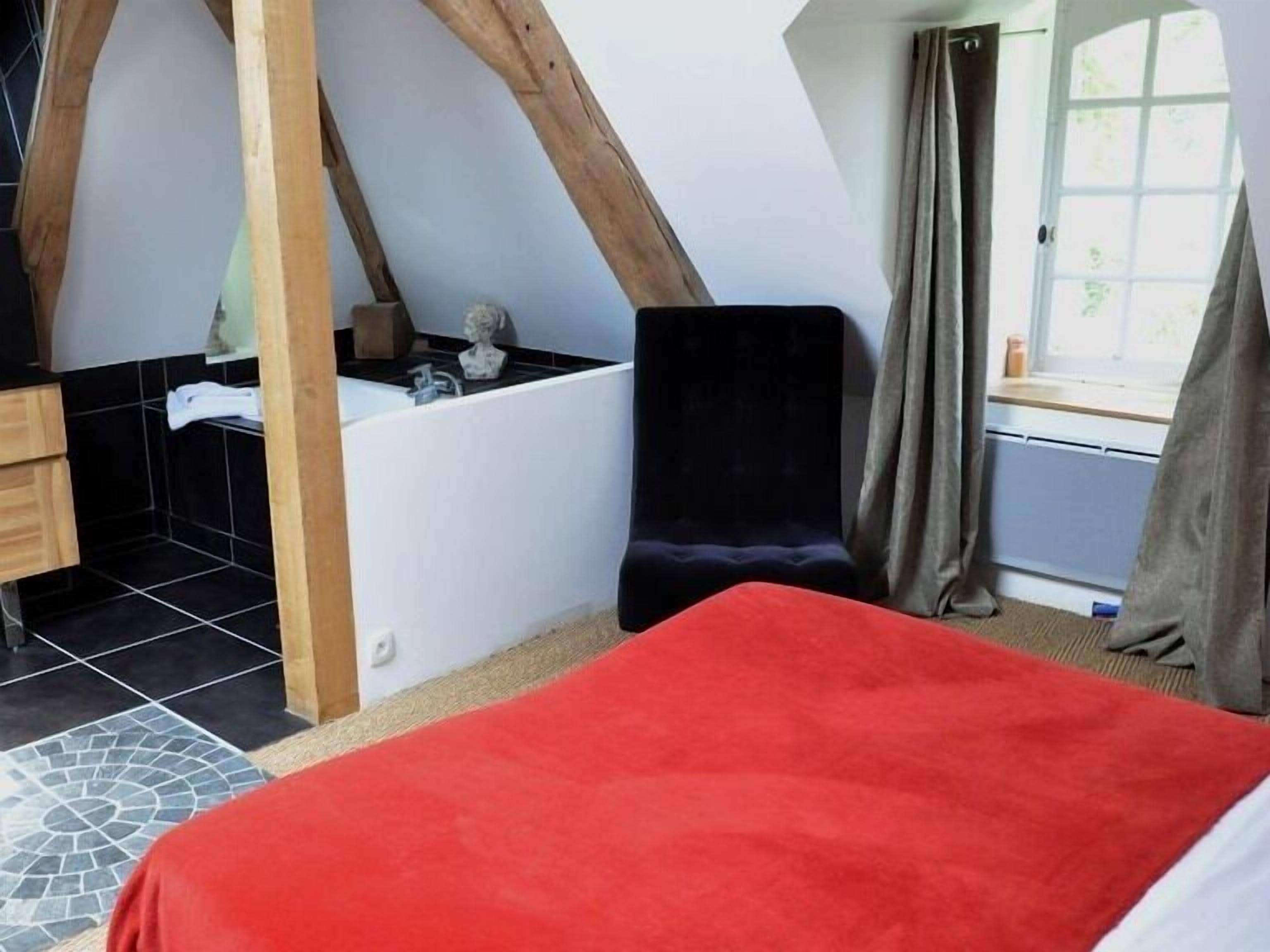 A bedroom with a sloped ceiling, a bathtub, a red bedspread, and a window with curtains.