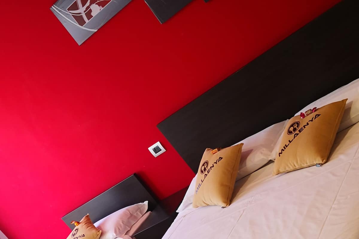 A hotel room with two beds, each with white bedding and pillows labeled 'MILLENYA'. The room has a red accent wall and a black headboard.