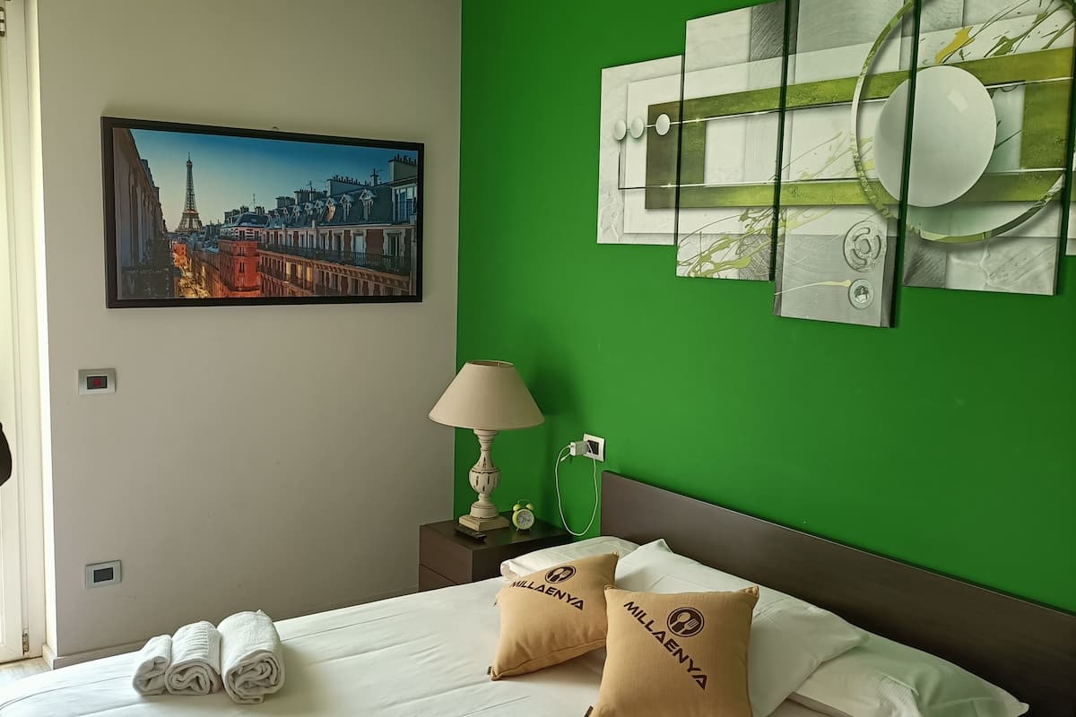 A hotel room with a bed, pillows, a nightstand with a lamp, a painting of a cityscape, and a green accent wall.