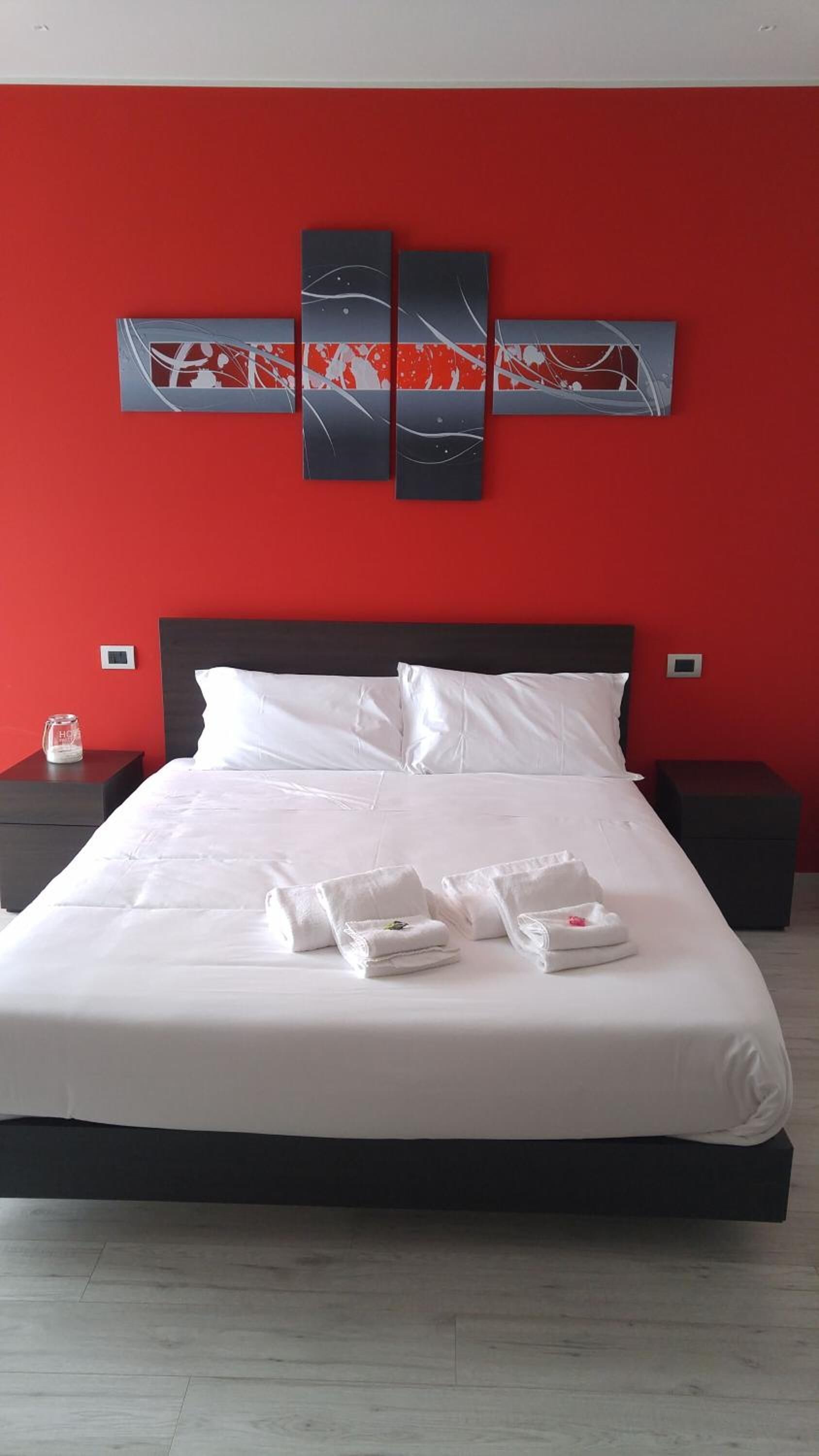 Quarto de hotel com uma cama grande, roupa de cama branca, parede de destaque vermelha e uma pintura abstrata moderna acima da cama.