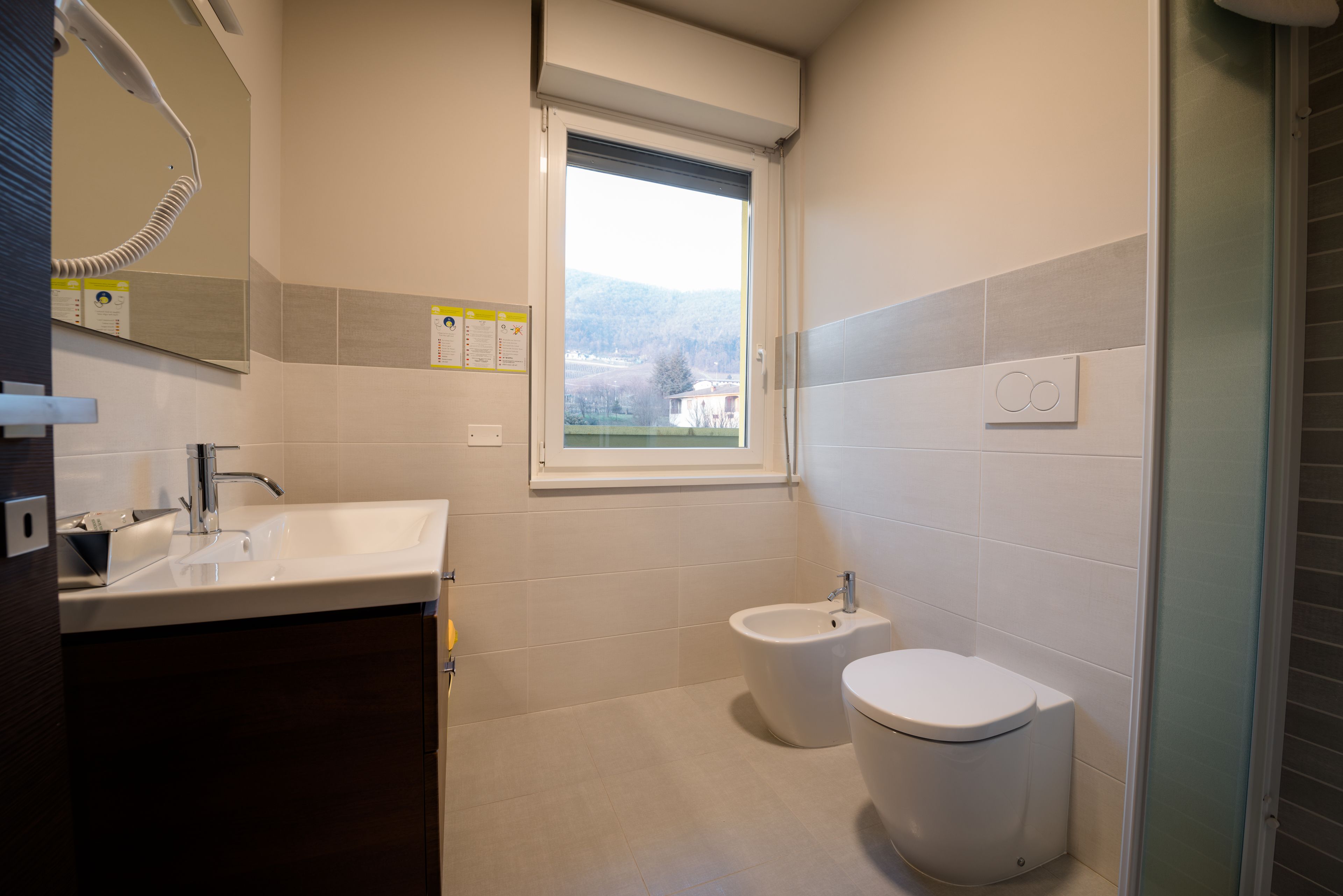 A modern bathroom with a sink, bidet, and toilet. There is a window providing natural light and a view of the outside.