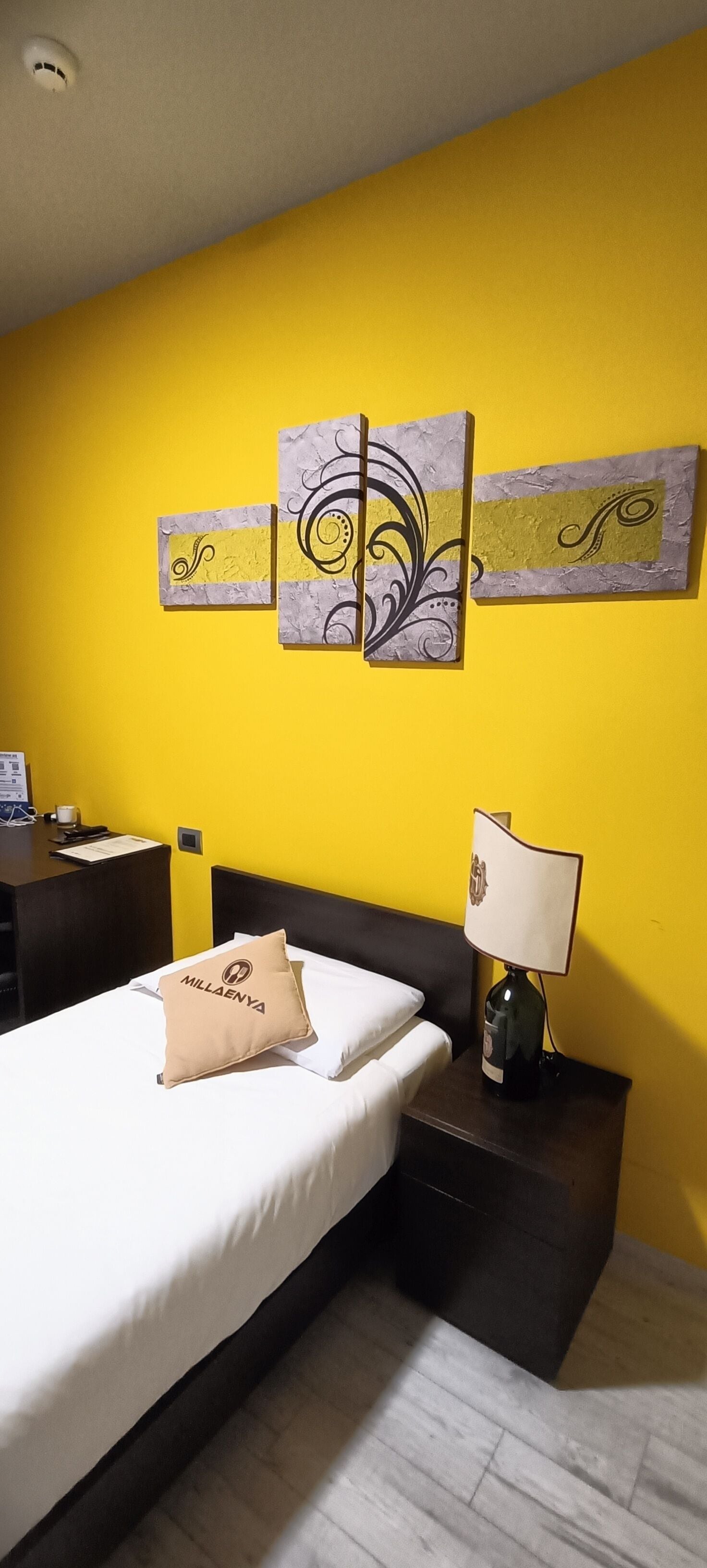 Ein Hotelzimmer mit gelber Wand, einem Bett mit einem Kissen mit der Aufschrift "MILLAENYA", einem Nachttisch mit einer Lampe und zwei abstrakten Wandbildern.