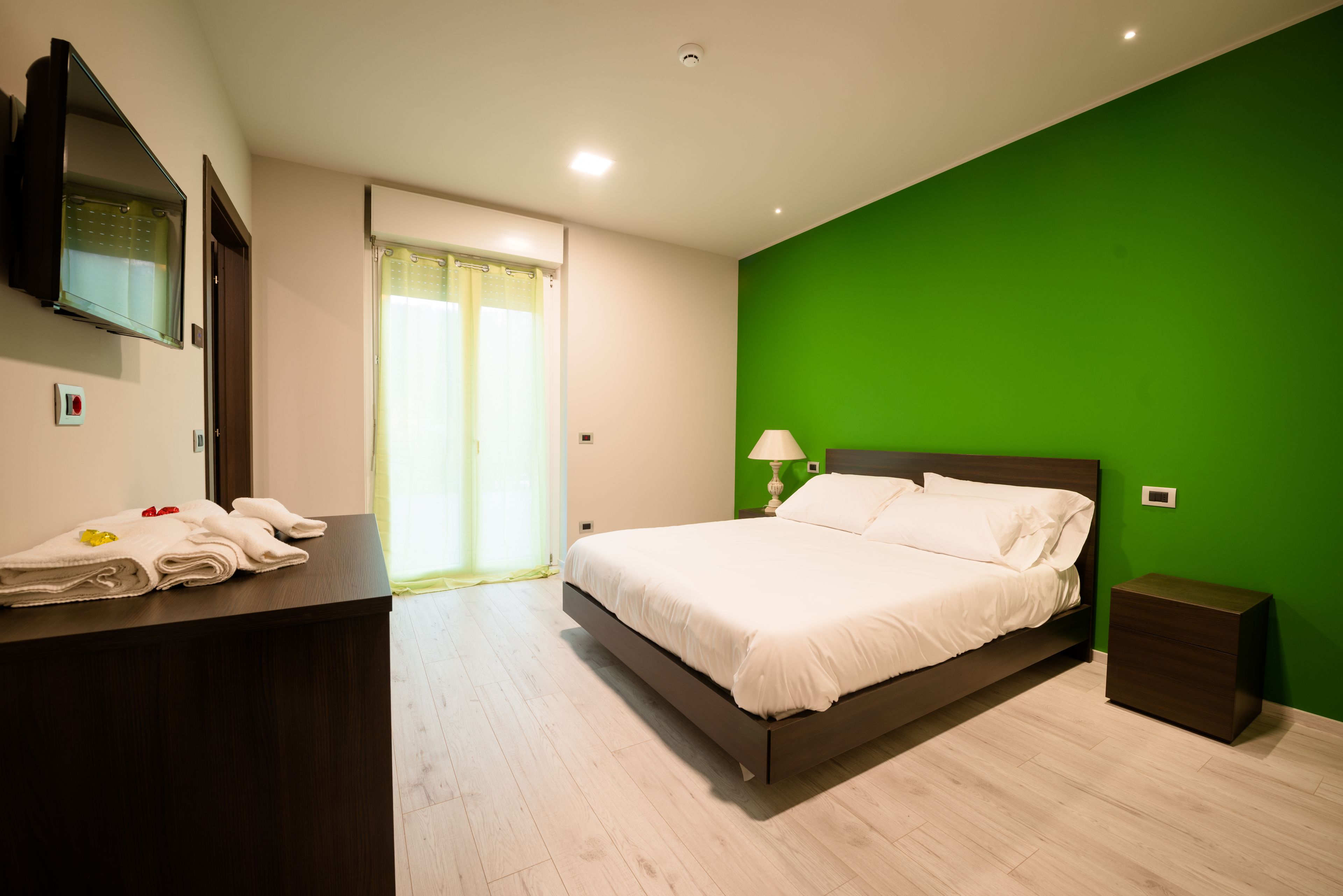 Un dormitorio con cama, mesita de noche, escritorio con toallas, televisor y pared verde.
