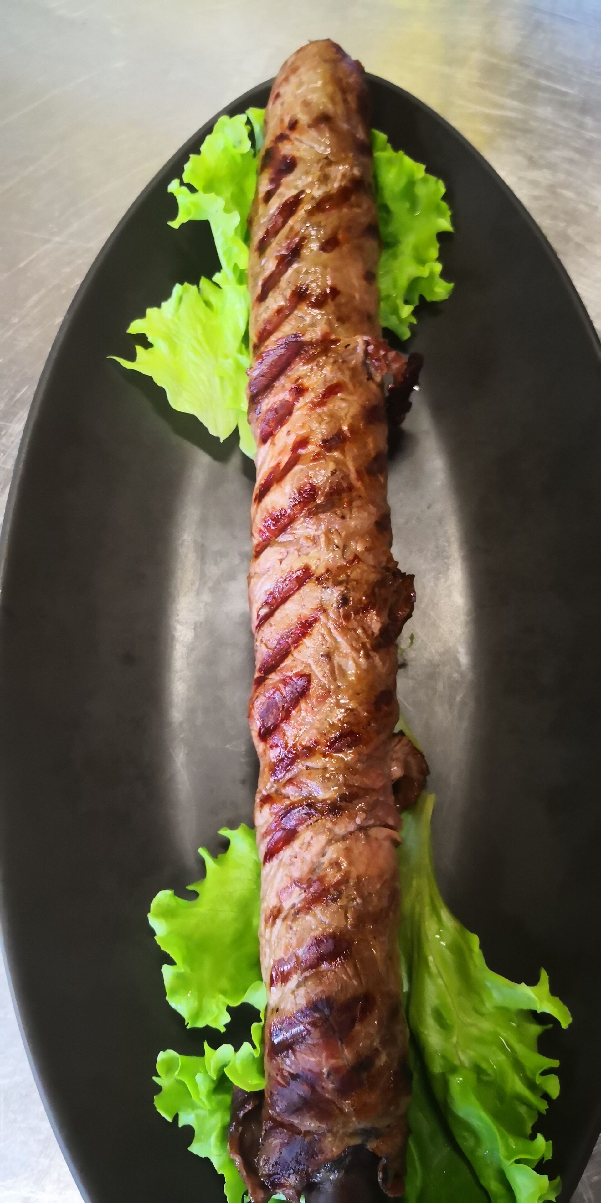 A long, grilled meat dish on a black plate with green lettuce leaves.
