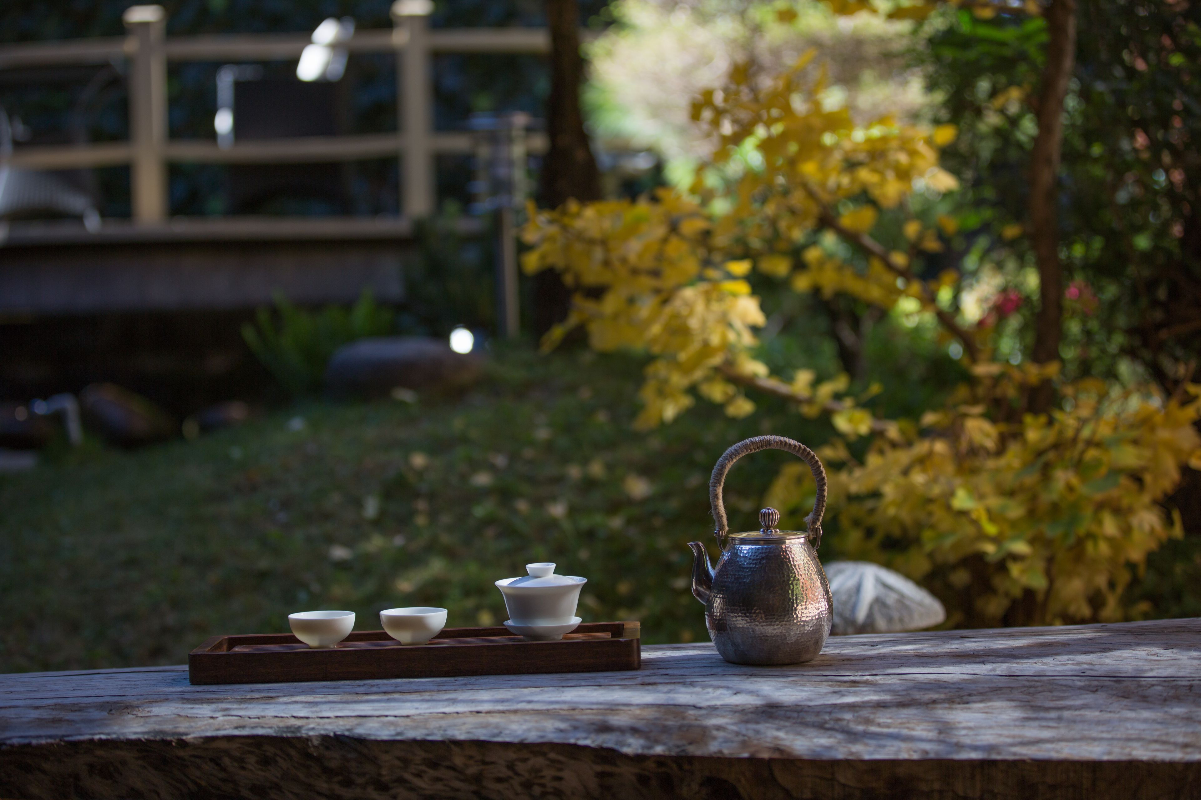 A rustic outdoor setting with a wooden table displaying a teapot and three cups.