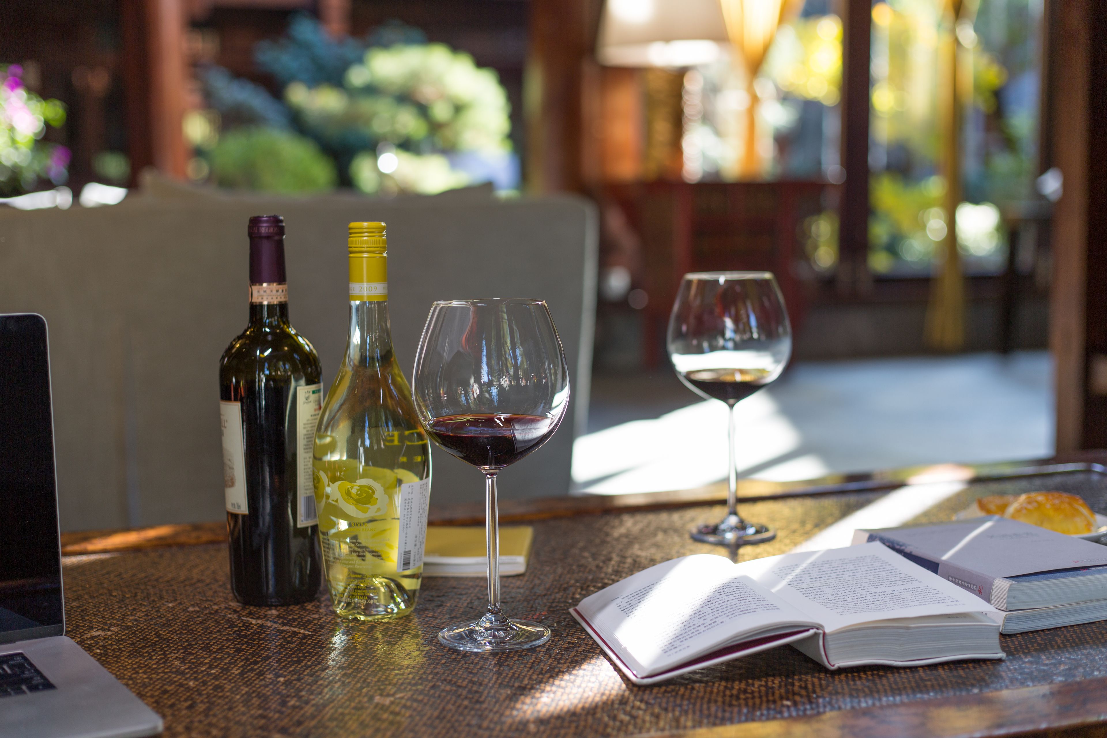 A table with two wine bottles, two wine glasses, a laptop, and an open book.