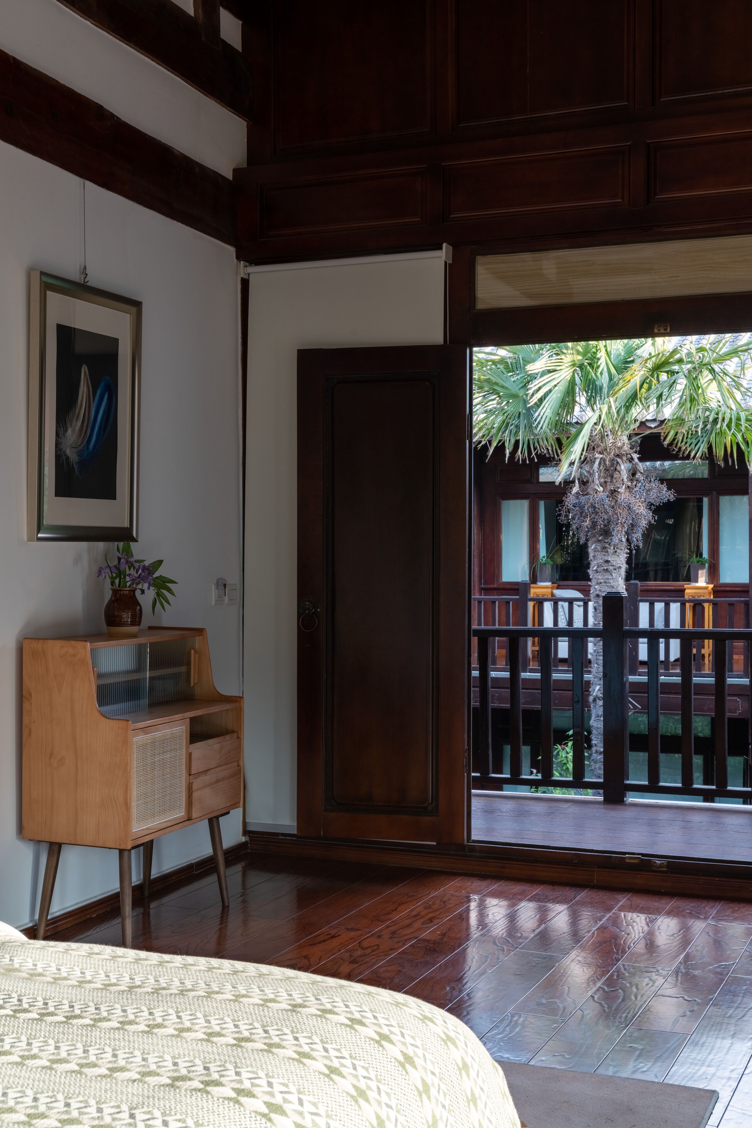 Une pièce comprenant un lit, une armoire en bois, un tableau encadré et une porte donnant sur un espace extérieur avec un palmier et une terrasse en bois.