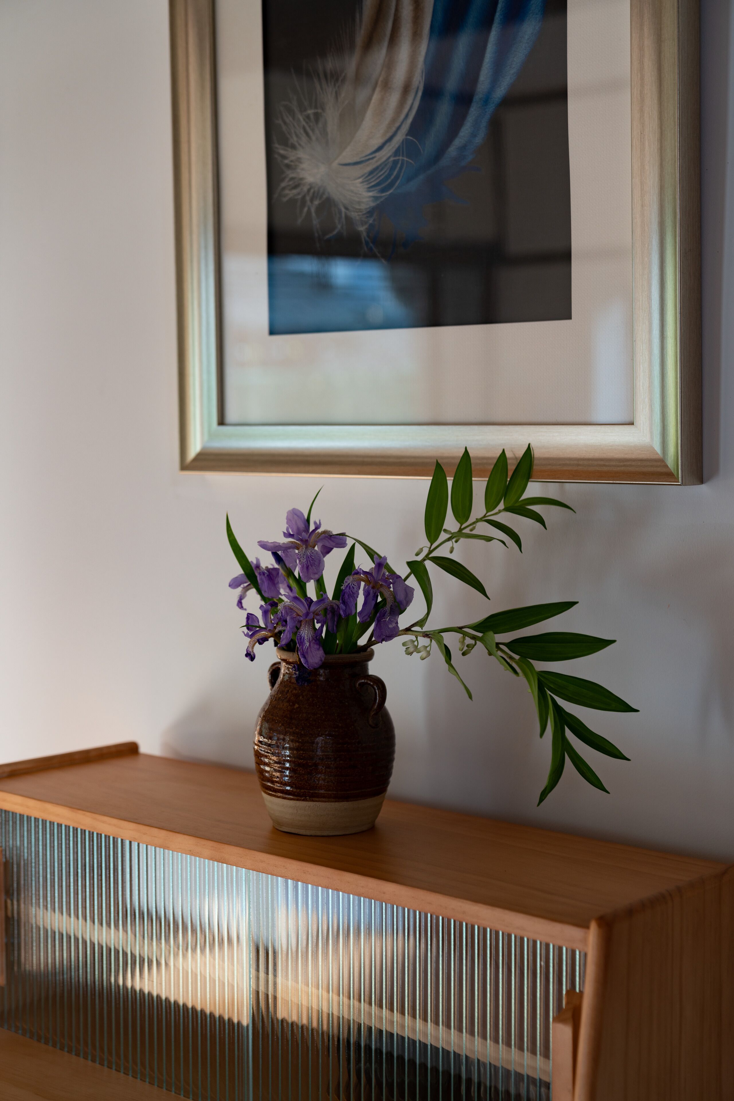 Un tableau encadré est accroché au-dessus d’une console en bois, sur laquelle se trouve un vase en céramique contenant des fleurs violettes et des feuilles vertes.