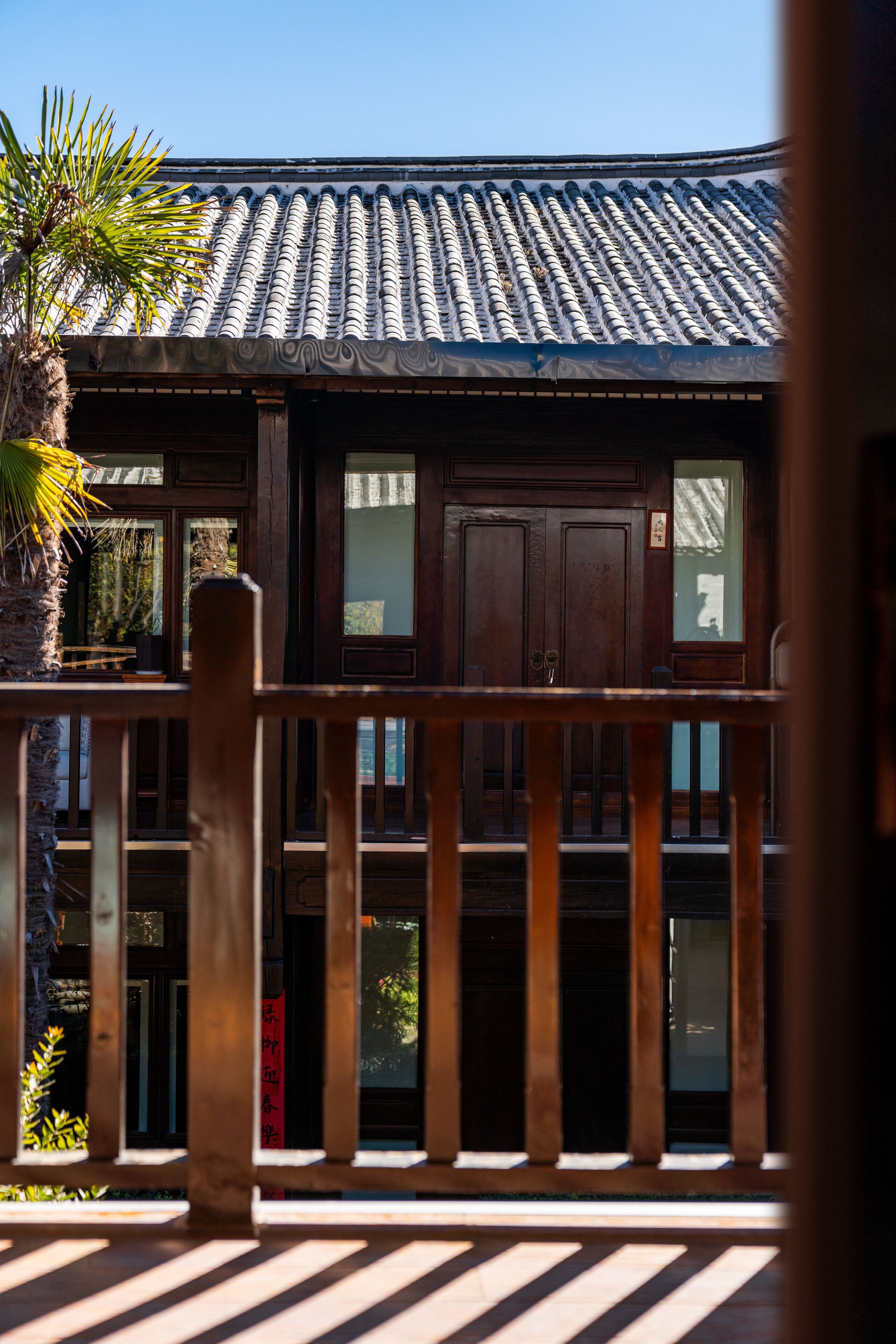 A traditional building with a tiled roof, wooden balcony railing, and a palm tree in the foreground.