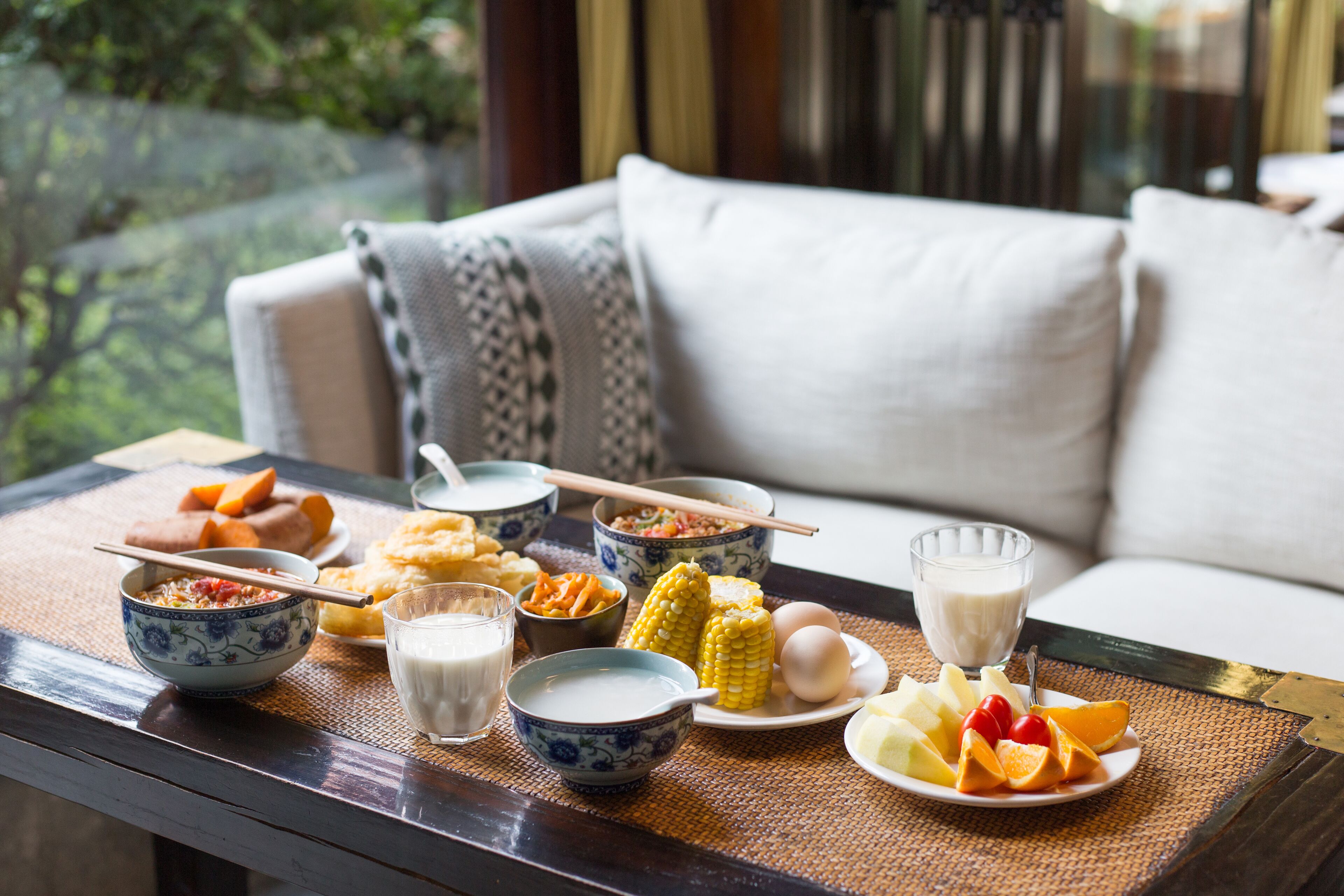 A dining table with various dishes, including bowls of soup, corn on the cob, and fruit, set on a woven mat.