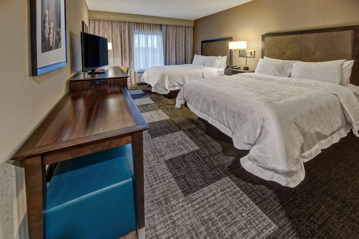 Une chambre d’hôtel avec deux lits, un bureau, une télévision et une grande fenêtre avec des rideaux.