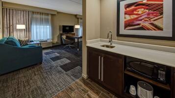 Habitación de hotel con sofá, escritorio, televisor y una kitchenette equipada con microondas, además de un cuadro en la pared.
