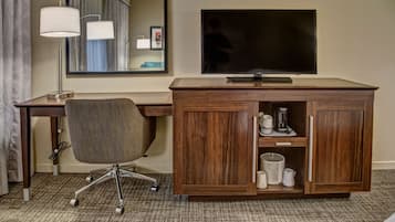A hotel room with a wooden desk, a modern chair, a flat-screen TV, and a cabinet with toiletries.