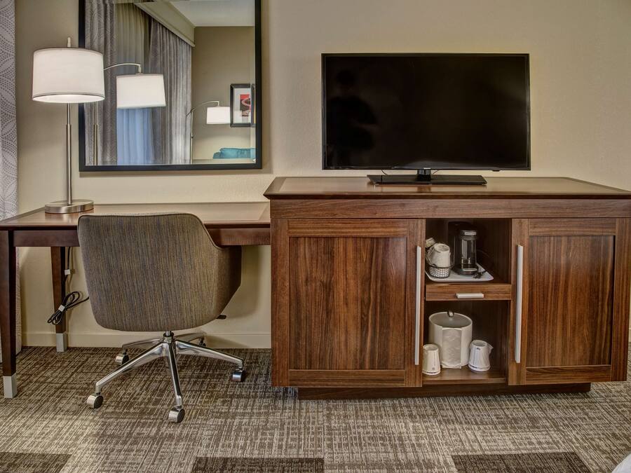 A hotel room with a wooden desk, a modern chair, a flat-screen TV, and a cabinet with toiletries.
