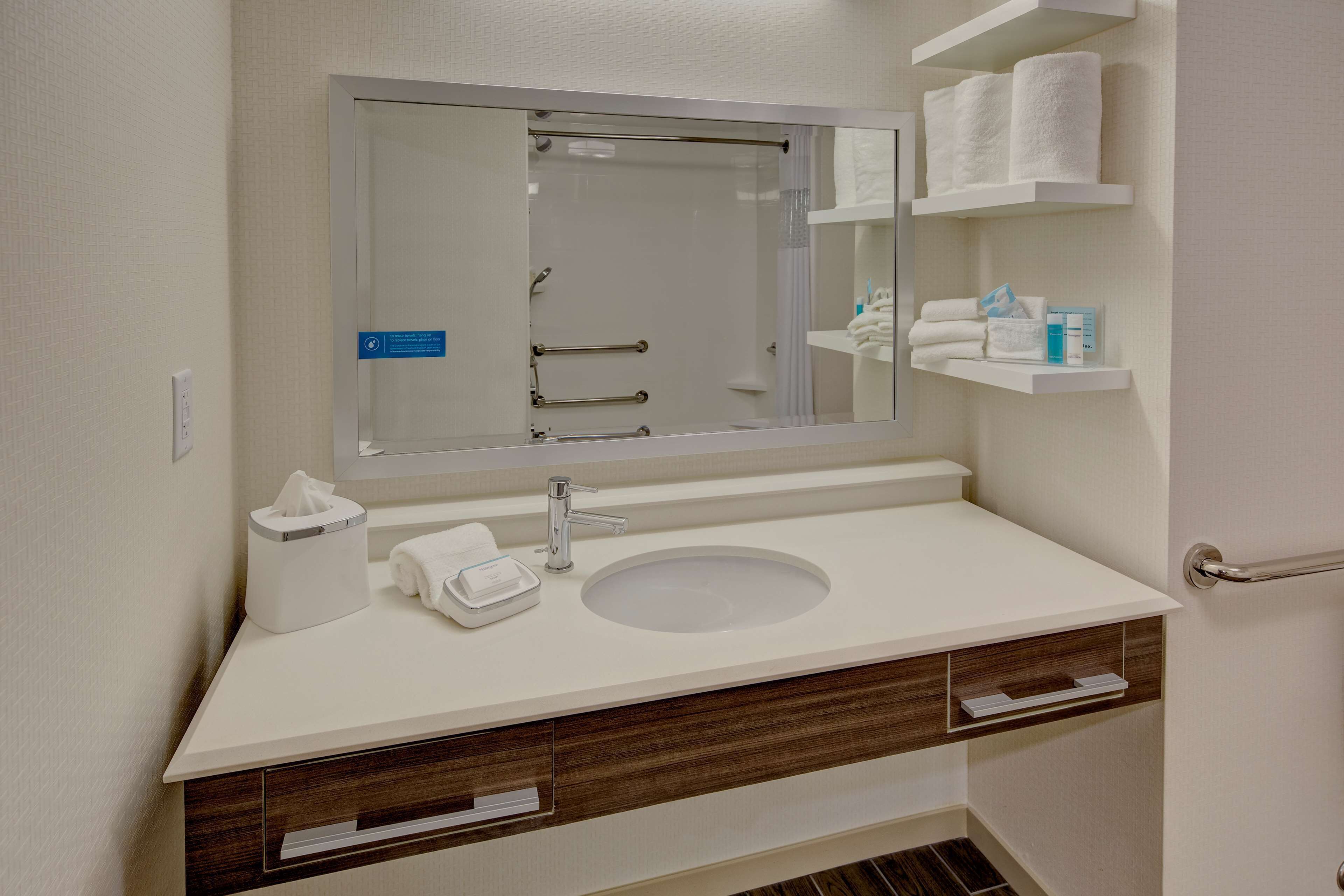 A modern bathroom with a large mirror, white sink, and wooden vanity. There are towels and toiletries on the shelves.