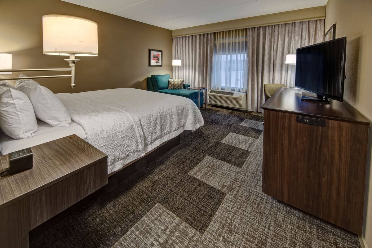 Une chambre d’hôtel avec un grand lit, un bureau avec une lampe, une télévision et une fenêtre avec des rideaux.