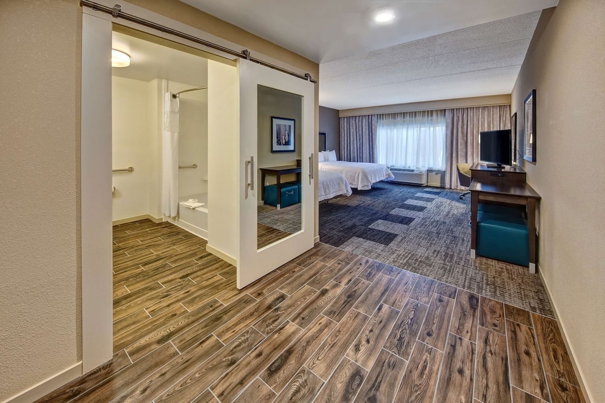 Une chambre d’hôtel avec un plancher en bois, un lit, un bureau, une télévision et une salle de bain équipée d’une douche et de toilettes.