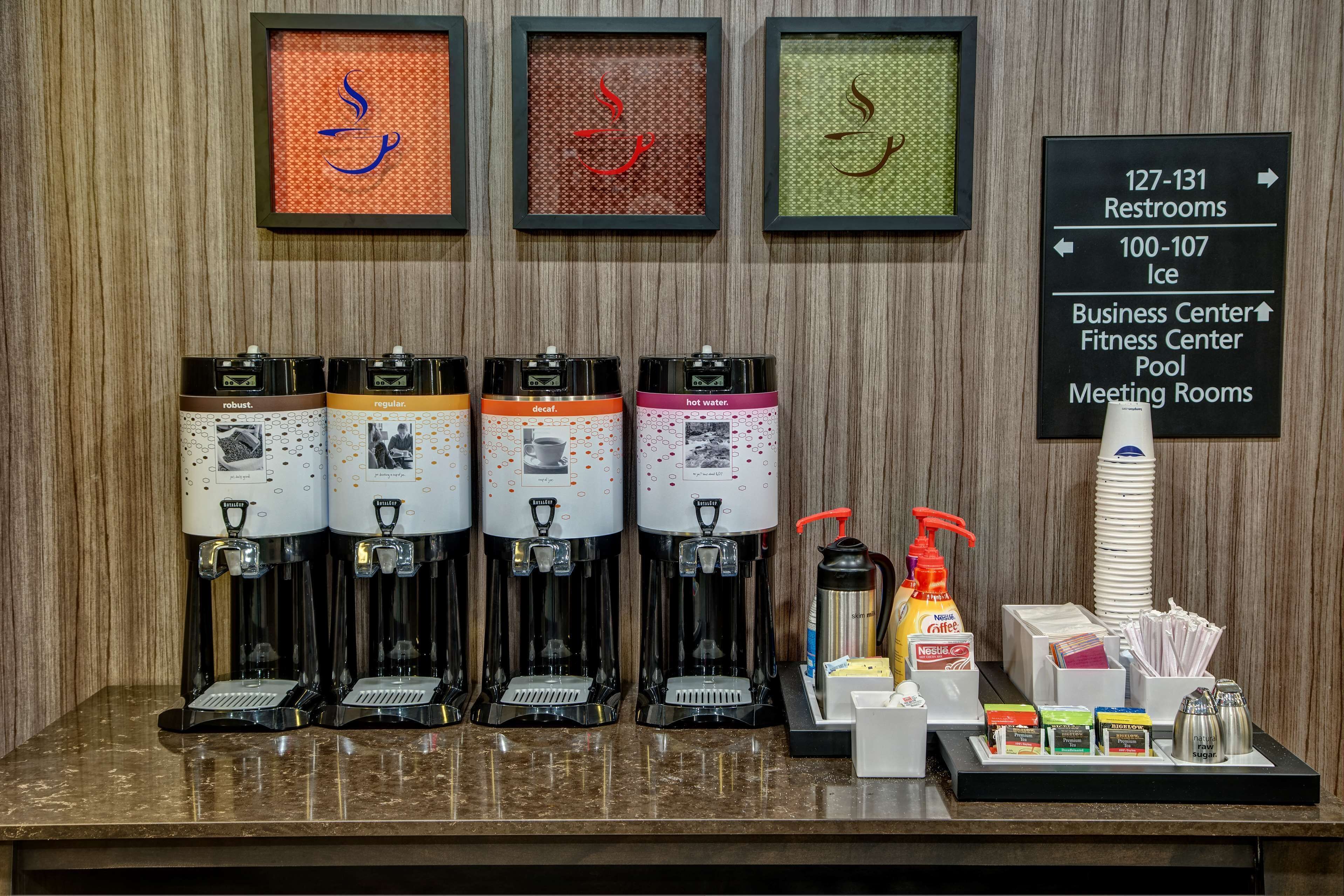 Estação de café com quatro dispensadores, uma máquina de café e um dispensador de água. Há placas indicando sanitários, gelo e salas de reunião.