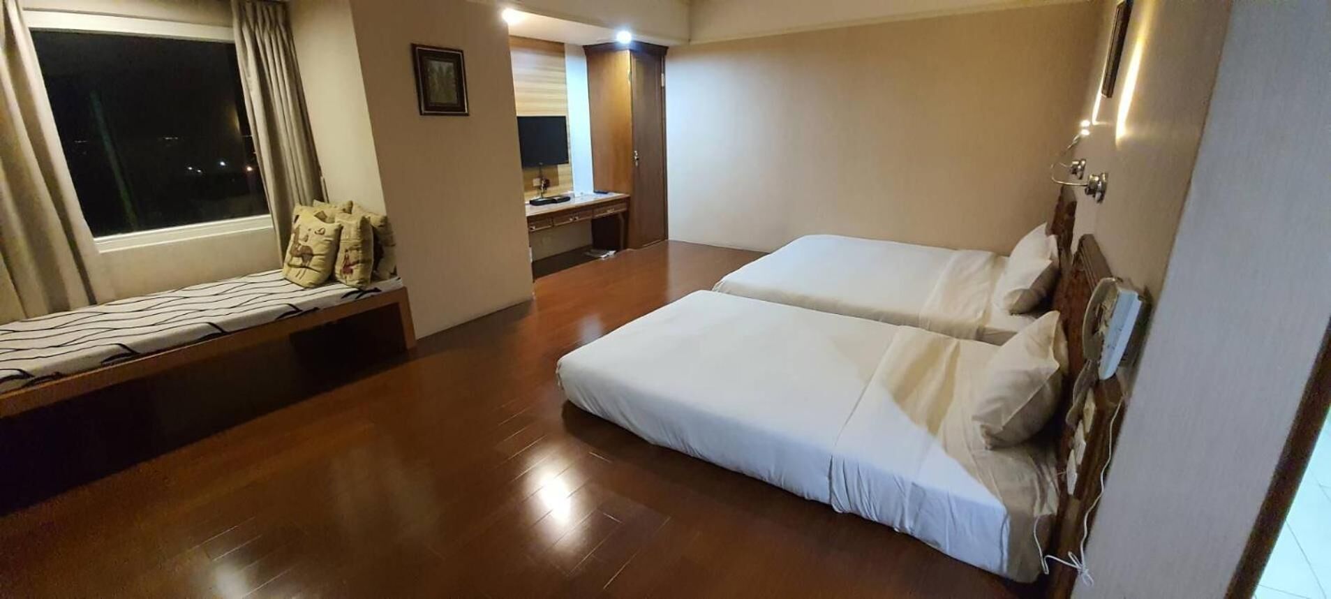 A hotel room with two beds, a wooden floor, a window with curtains, a bedside table, and a small desk with a chair.