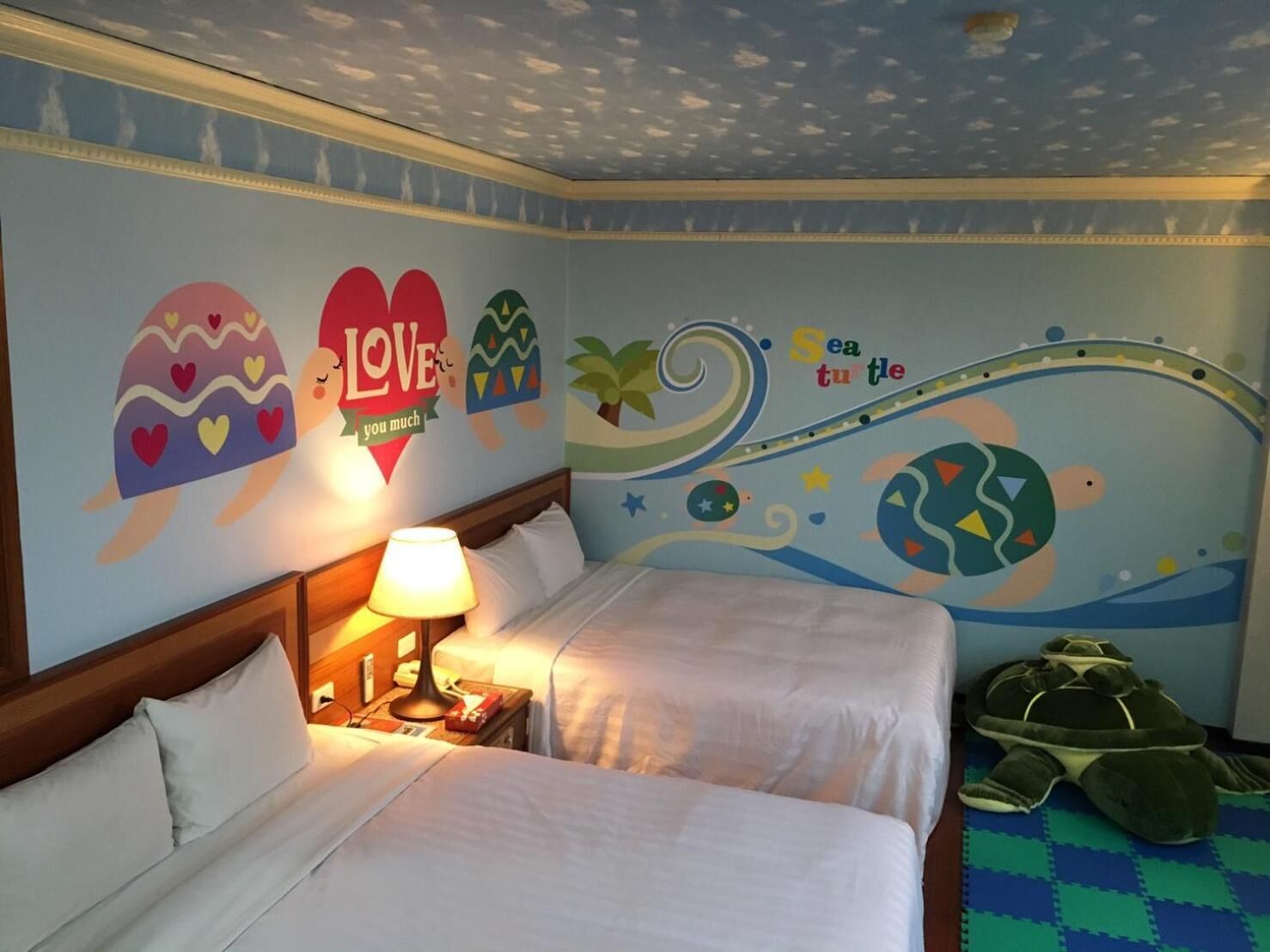Quarto de hotel com duas camas, uma mesa de cabeceira com abajur e um brinquedo de pelúcia em formato de tartaruga no chão.