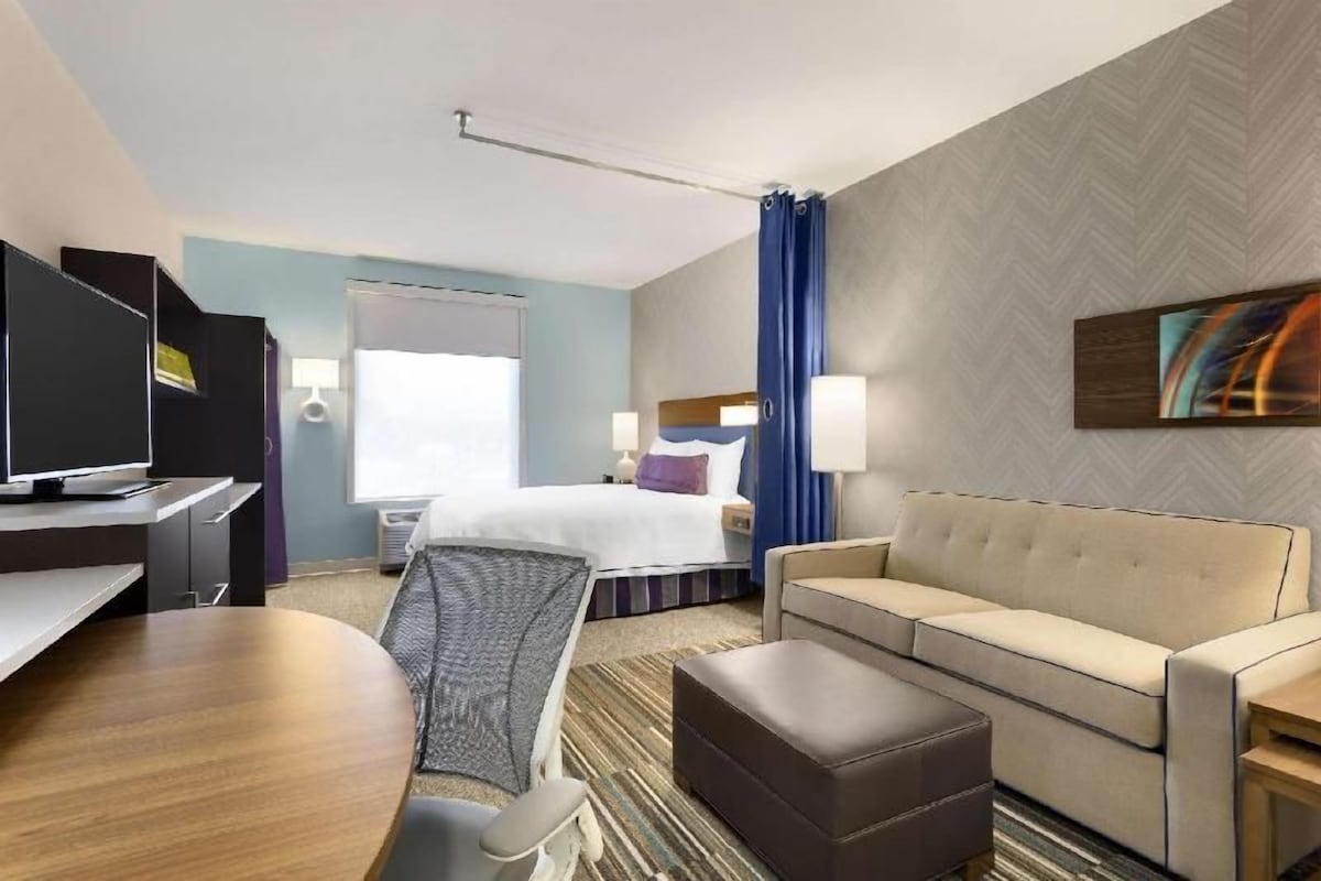 A hotel room with a bed, a sofa, a TV, a desk, and a window with curtains.