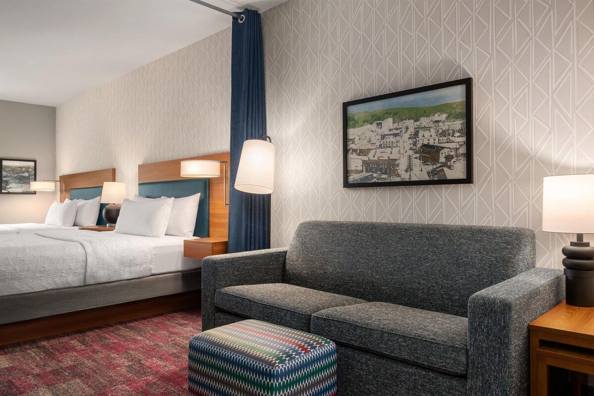 A hotel room with a bed, a sofa, a patterned ottoman, a painting, and a patterned wall.