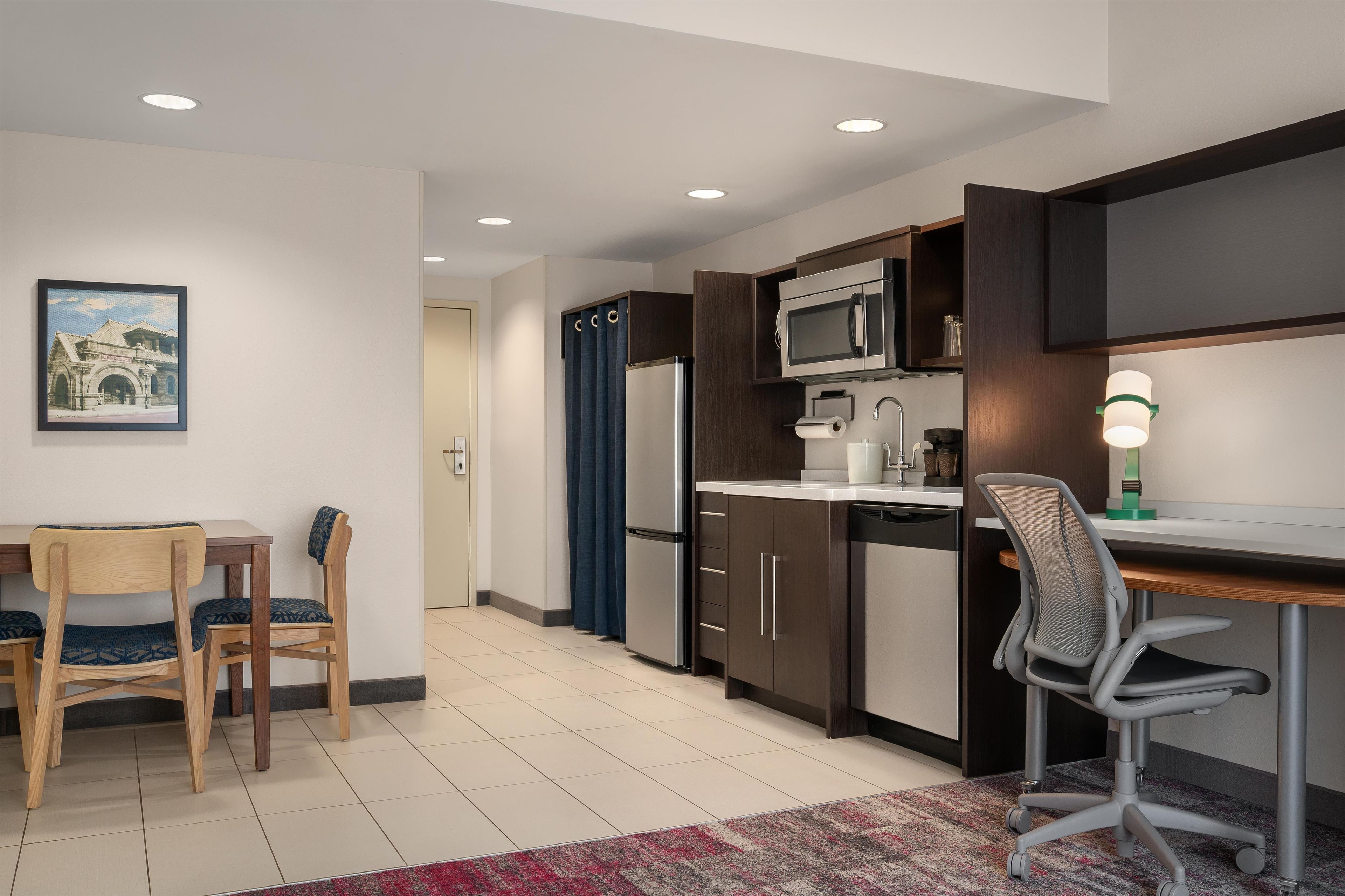A modern kitchen with dark cabinetry, a microwave, and a refrigerator. There is a dining area with a table and chairs, and a framed picture on the wall.