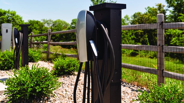 Una estación de carga para vehículos eléctricos negros, con cables conectados, frente a una cerca de madera y follaje.