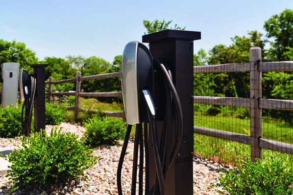 A black electric vehicle charging station with cables connected to it, set against a wooden fence and greenery. - Home2 Suites by Hilton Middletown (Middletown)