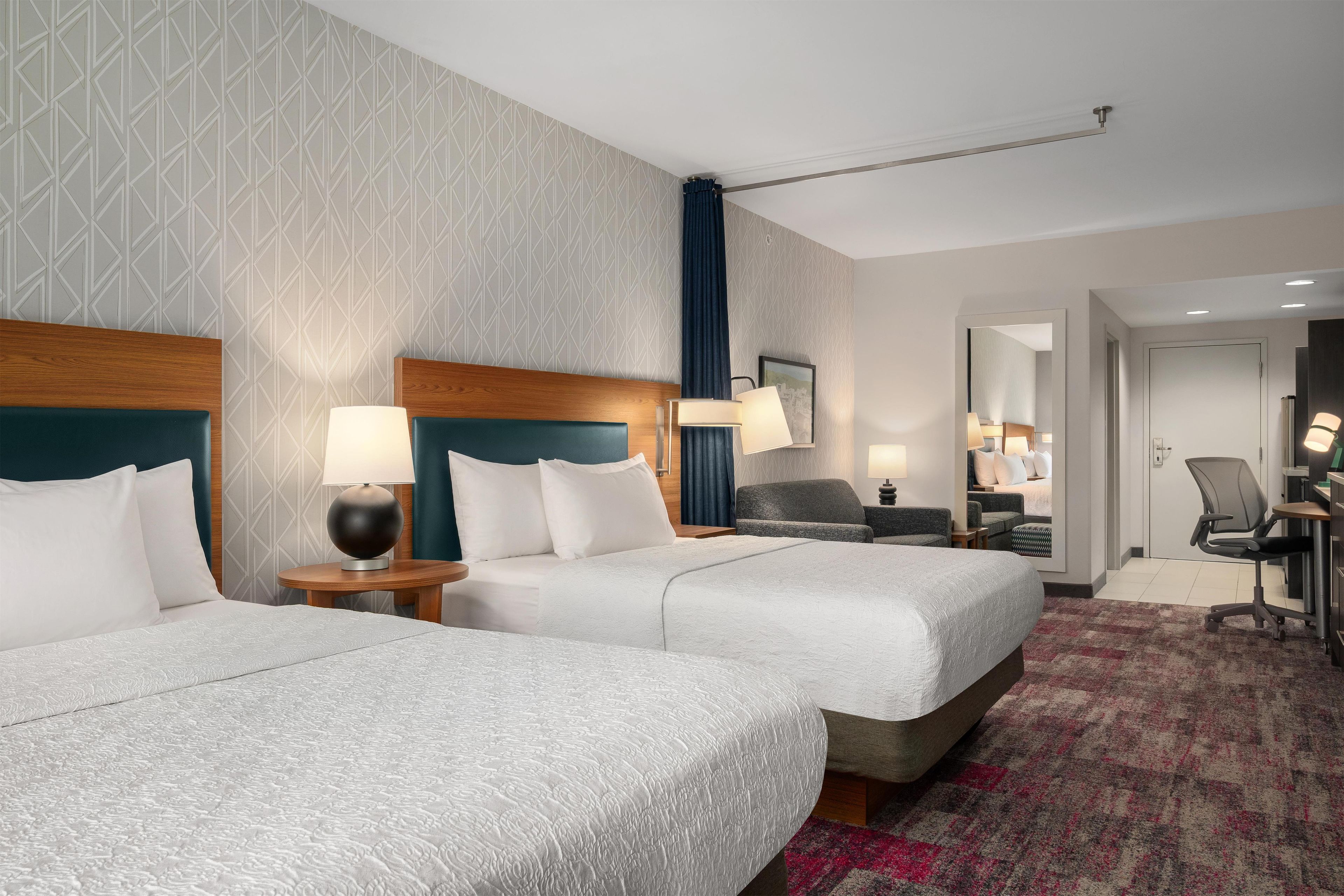 A hotel room with two beds, a desk, a chair, and a bathroom visible through an open door.