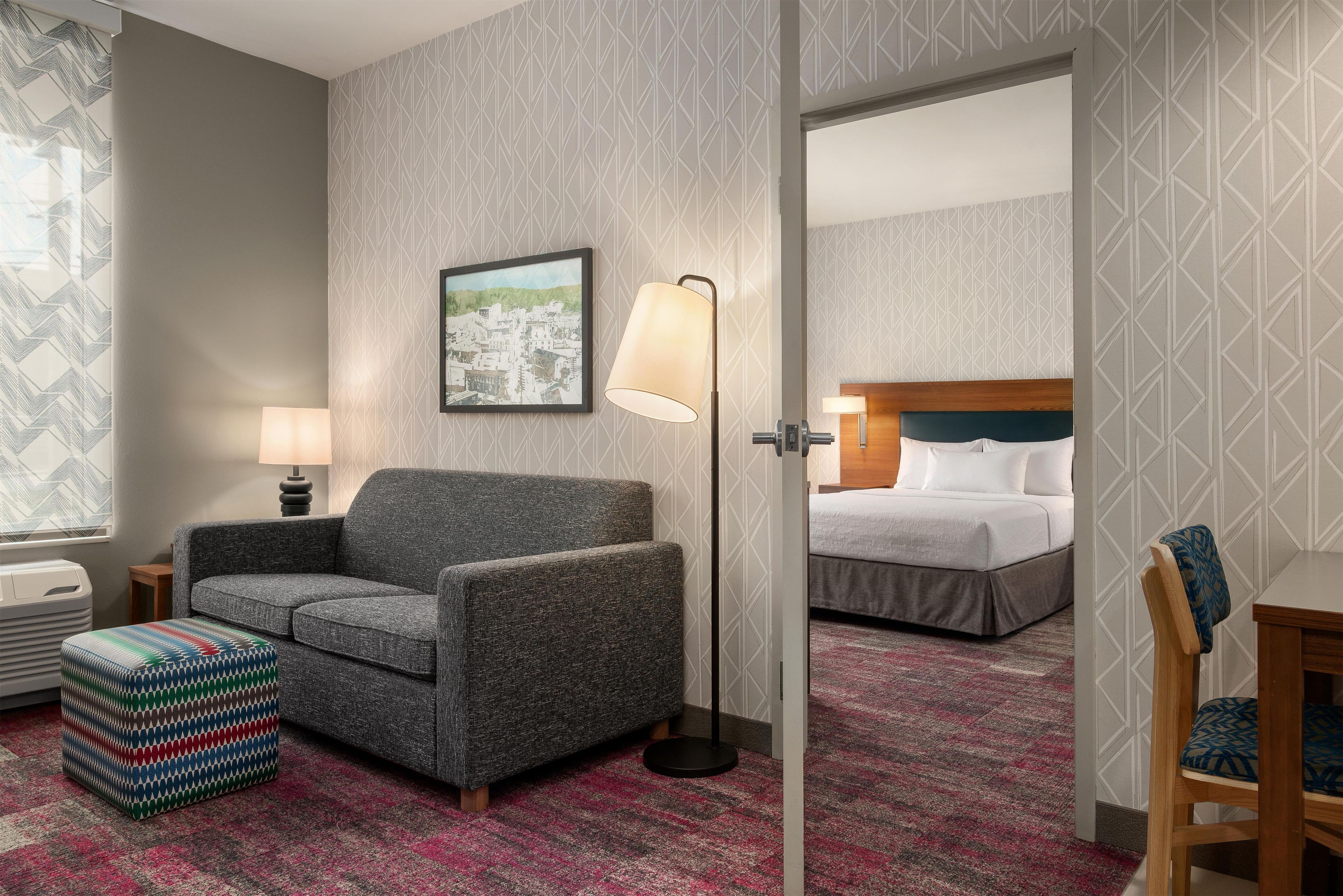 A hotel room with a gray armchair, a colorful ottoman, a framed cityscape picture, a standing floor lamp, and a bed with white linens.