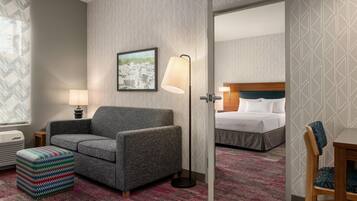 A hotel room with a gray armchair, a colorful ottoman, a framed cityscape picture, a standing floor lamp, and a bed with white linens.
