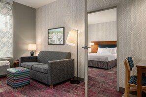 A hotel room with a gray armchair, a colorful ottoman, a framed cityscape picture, a standing floor lamp, and a bed with white linens. - Home2 Suites by Hilton Middletown (Middletown)