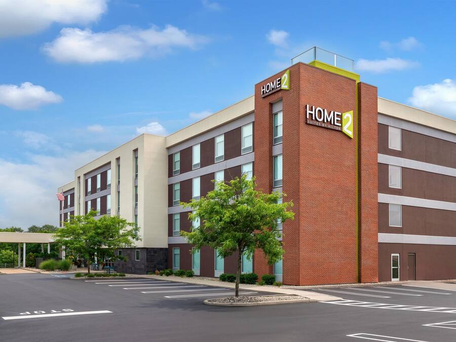 Un edificio de hotel de varios pisos con un llamativo letrero 'HOME 2', que presenta un diseño moderno con fachadas de ladrillo y color beige, y un estacionamiento al frente.