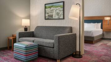 Una habitación de hotel con un sofá gris, un otomano colorido, un cuadro enmarcado de un paisaje urbano, una cama con ropa de cama blanca y dos lámparas de noche.
