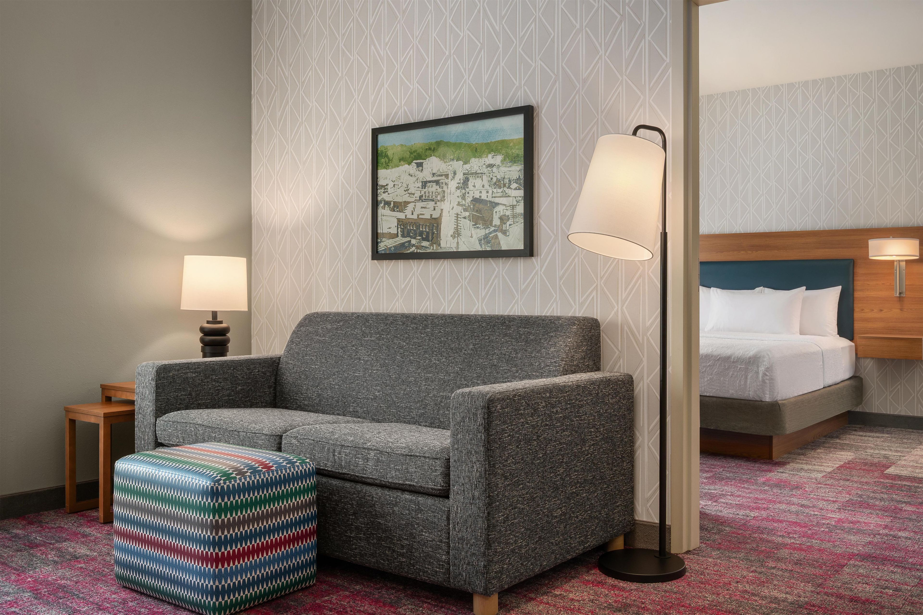 A hotel room with a gray sofa, a colorful ottoman, a framed cityscape painting, a bed with white bedding, and two bedside lamps.