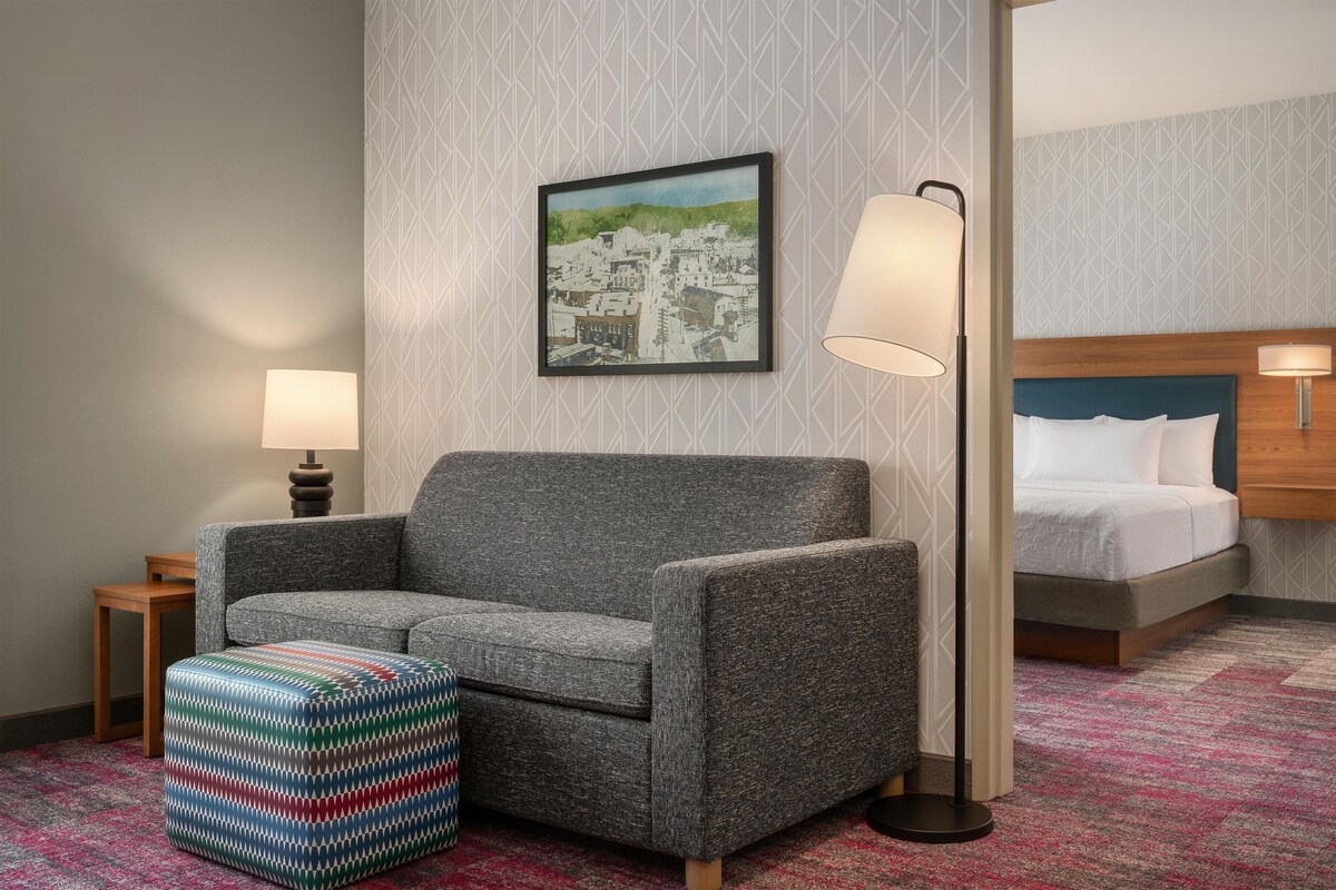 A hotel room with a gray sofa, a colorful ottoman, a framed cityscape painting, a bed with white bedding, and two bedside lamps.