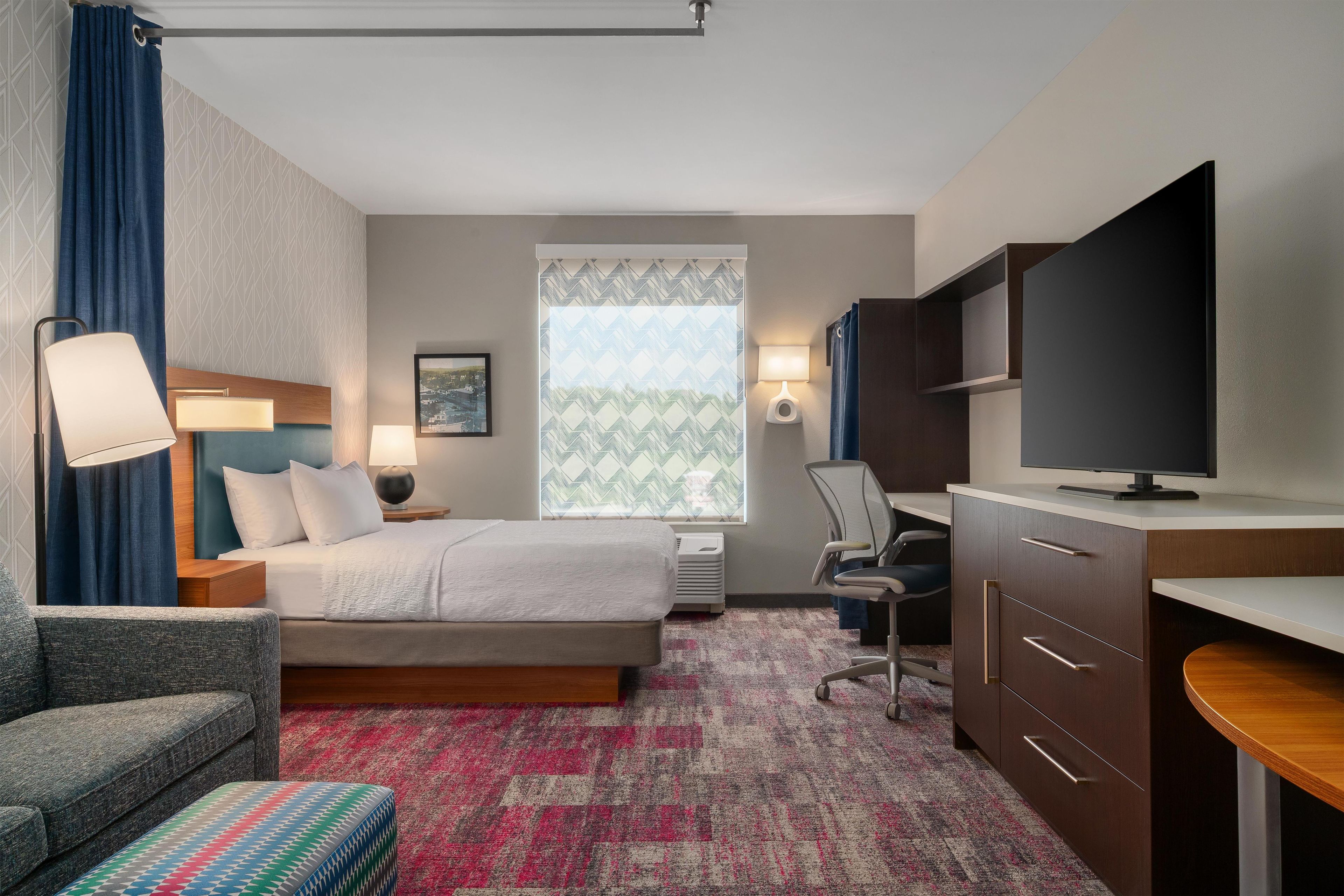 A hotel room with a bed, a desk, a television, a chair, a sofa, and a window with patterned curtains.