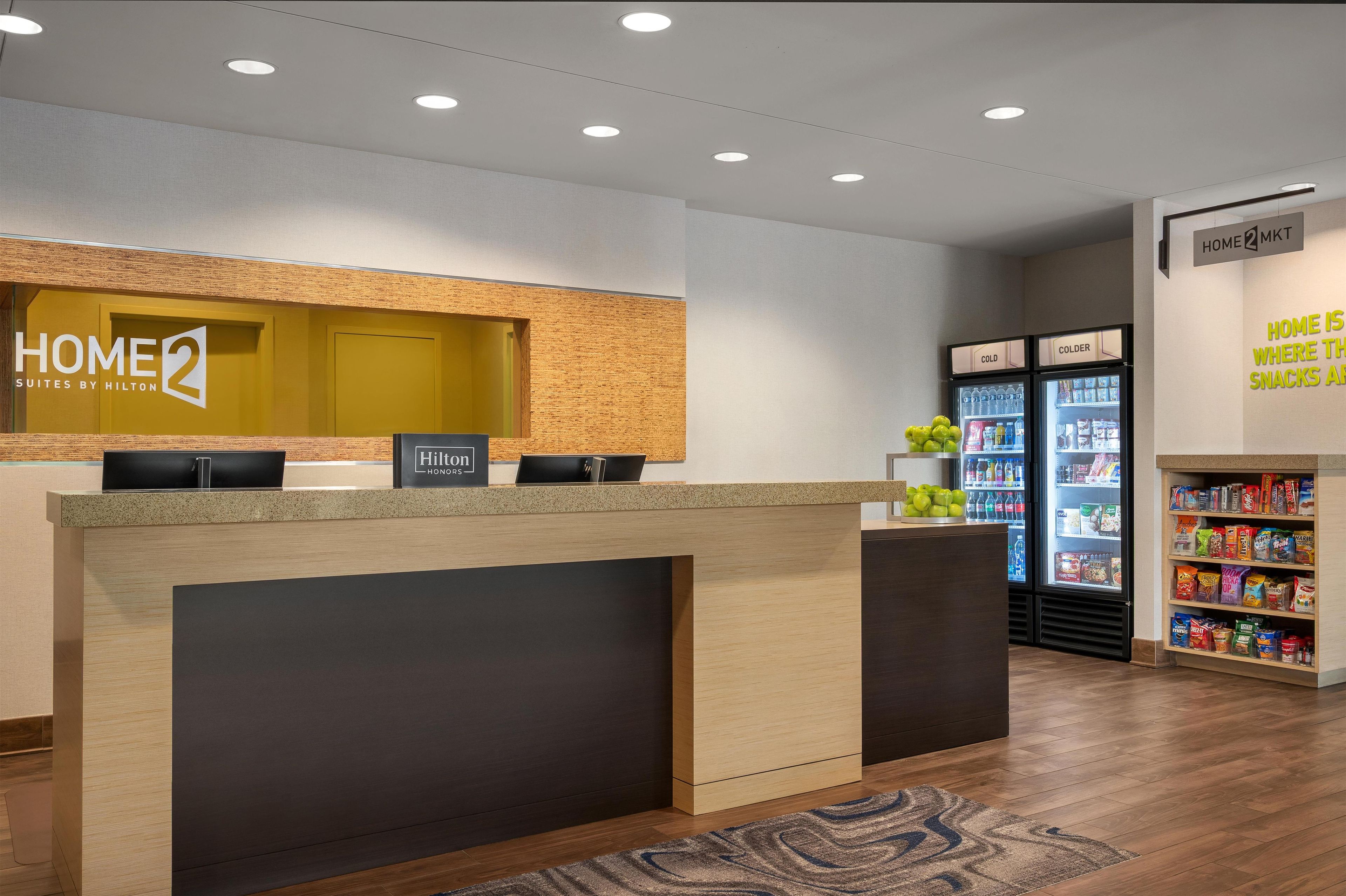 Zona de recepción de un hotel con mostrador, un cartel que dice 'Home 2 Suites by Hilton' y un refrigerador lleno de refrigerios.