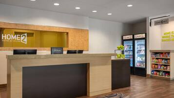 Zona de recepción de un hotel con mostrador, un cartel que dice 'Home 2 Suites by Hilton' y un refrigerador lleno de refrigerios.