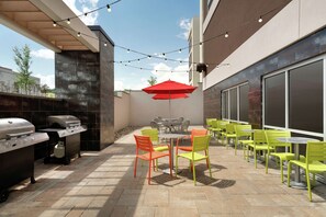 An outdoor patio with a grill, chairs, and tables, featuring a red umbrella and string lights. - Home2 Suites by Hilton Middletown (Middletown)