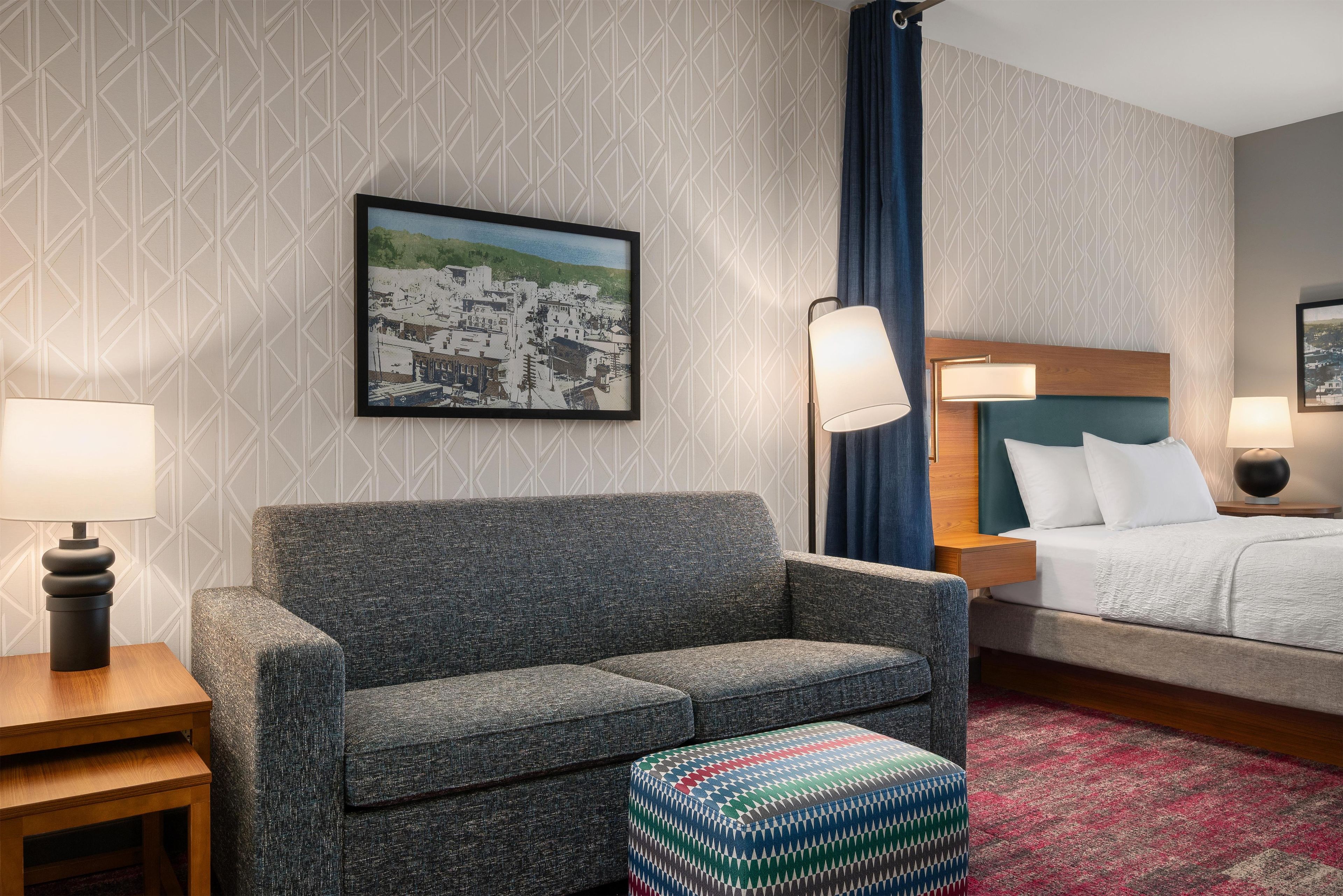 A hotel room with a sofa, a bed, a nightstand, a lamp, a picture on the wall, and a patterned rug.