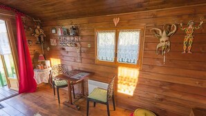 Ein gemütliches Holzhäuschen im Inneren mit einer Essecke, einem Tisch, Stühlen, einem Fenster mit Spitzenausschnitten und verschiedenen Wanddekorationen.