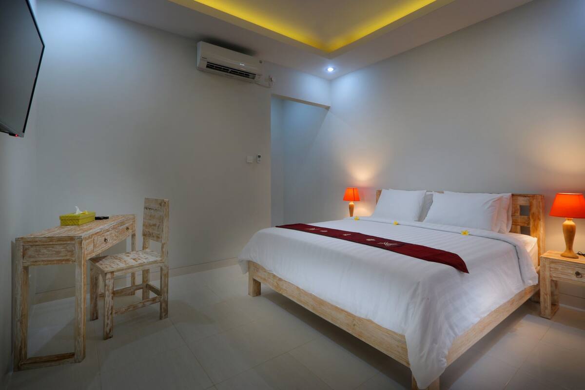 A bedroom with a wooden bed, two bedside lamps, a desk with a chair, and a TV mounted on the wall.