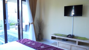 Une chambre d’hôtel comprenant un lit, un téléviseur fixé au mur, un petit meuble en bois servant de support pour le téléviseur, avec une télécommande, et une porte-fenêtre coulissante donnant sur un espace extérieur.