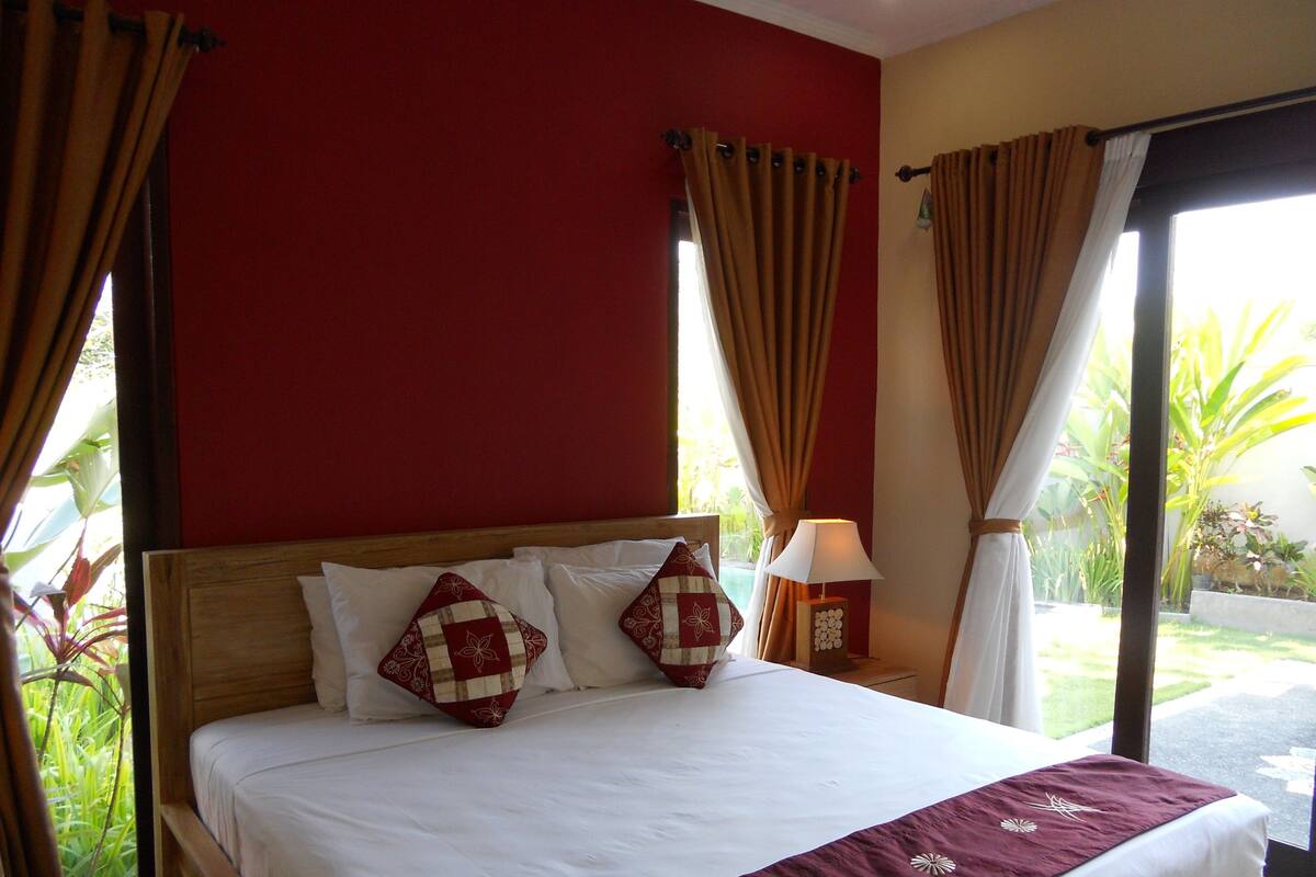 A hotel room with a bed, curtains, a nightstand with a lamp, and a view of greenery outside.