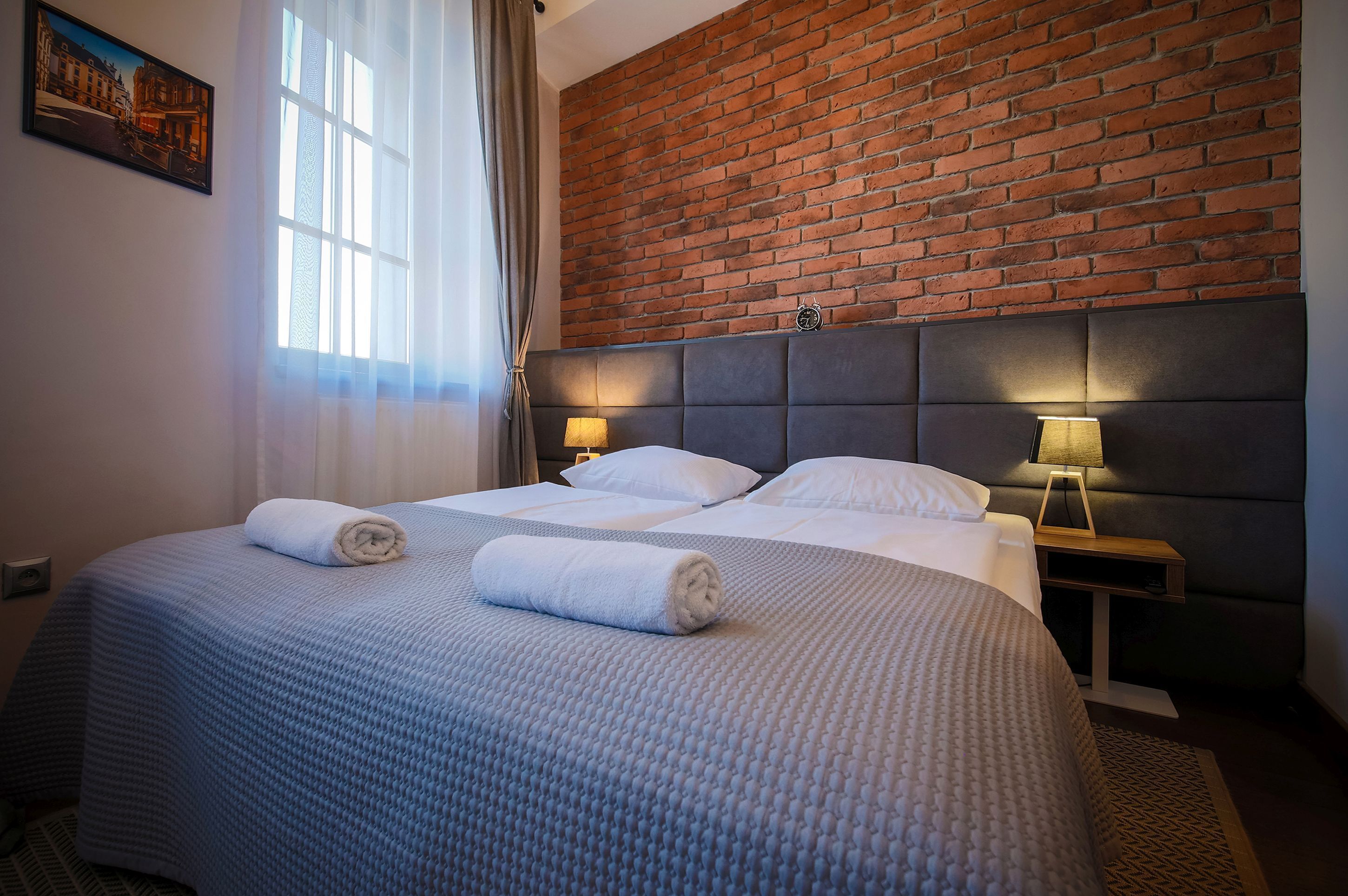 A hotel room with a brick wall, a large bed with two pillows, a bedside table with a lamp, and a window with curtains.